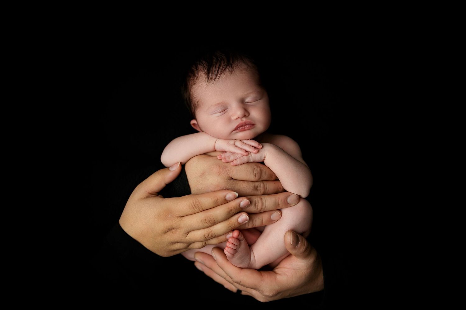 Artistic portrait of newborn cradled in hands against black background.