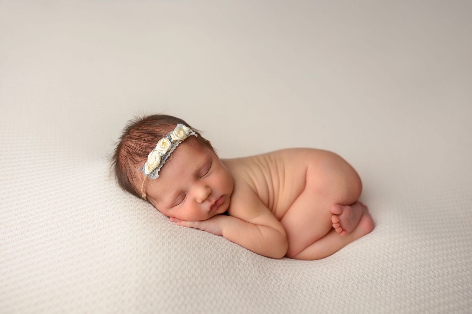 Sleeping newborn with headband curled up on white textured backdrop.