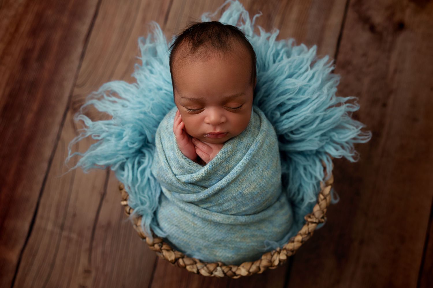 Smiling baby wrapped in turquoise fabric nestled in basket with fluffy blue backdrop.
