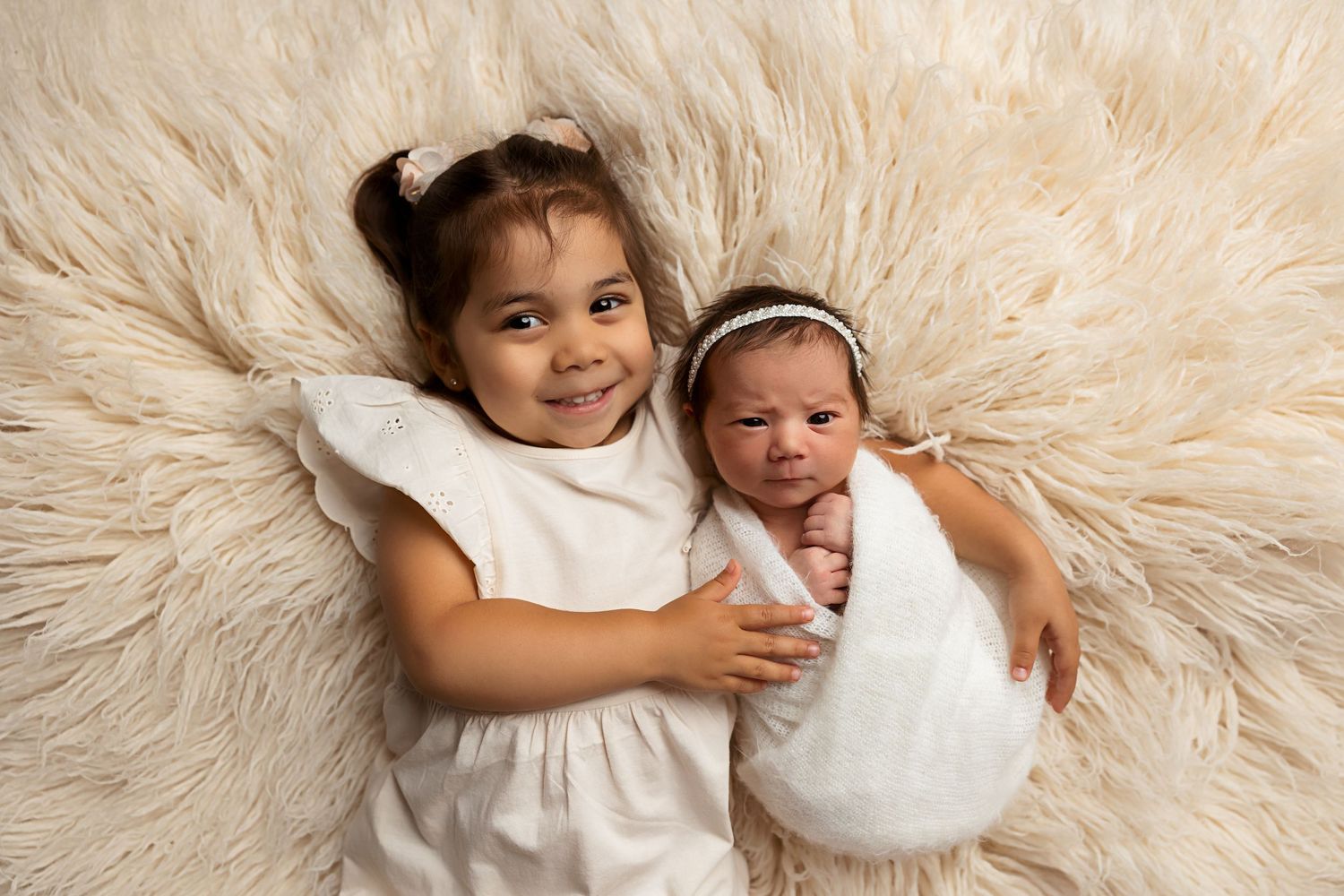 Big sister in white dress cuddling with newborn on soft furry backdrop.