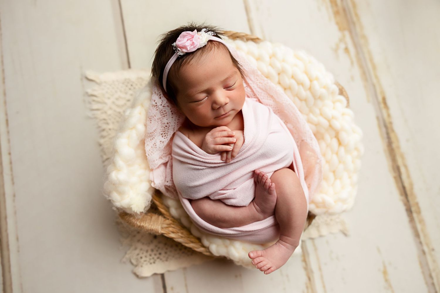 Newborn in pink wrap sleeping peacefully in rustic basket with cream fur lining.
