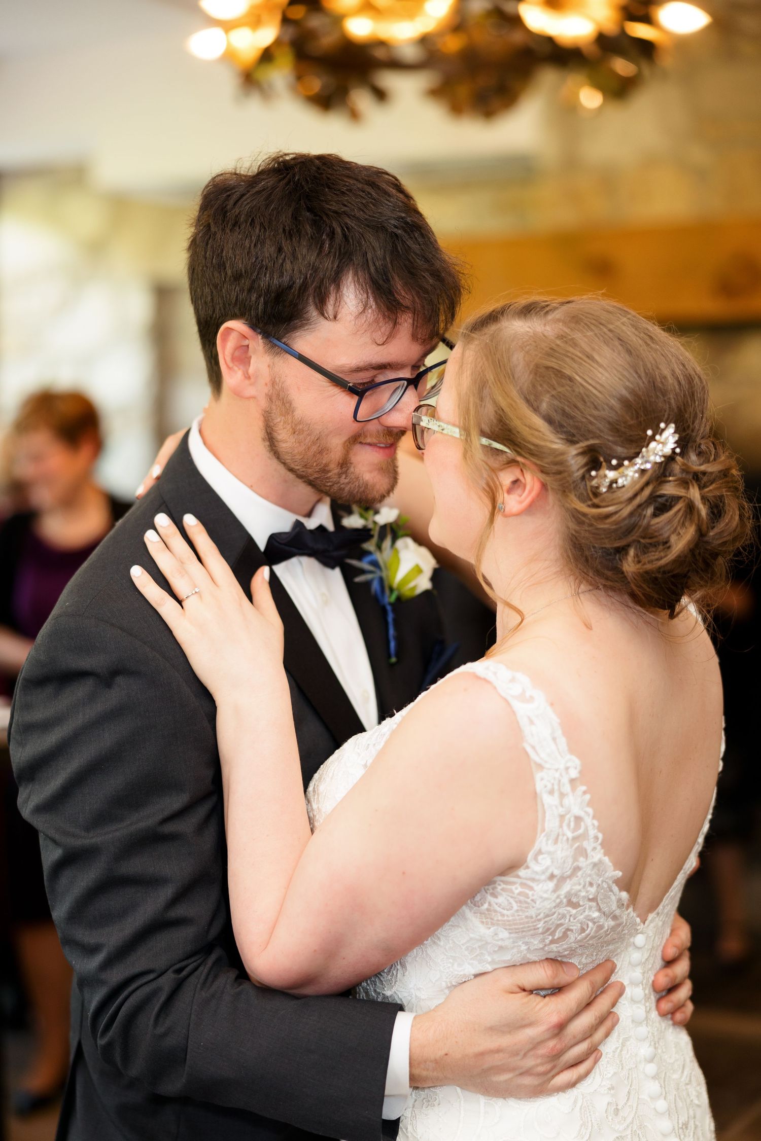 Romantic moment at wedding reception with elegant lace dress and formal attire under chandelier lighting.
