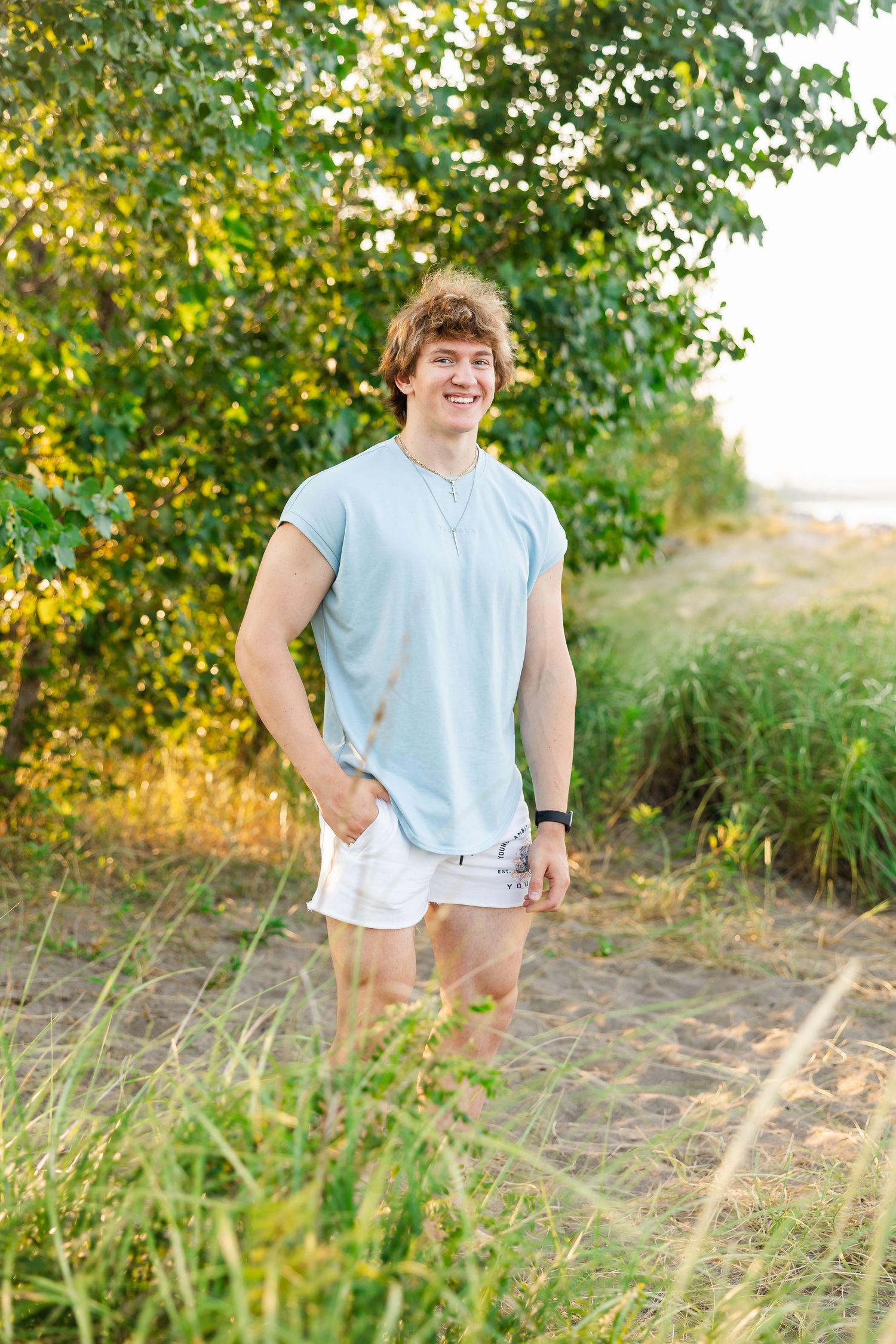 Someone poses with hands in pockets on a dirt path surrounded by autumn foliage and golden sunlight.