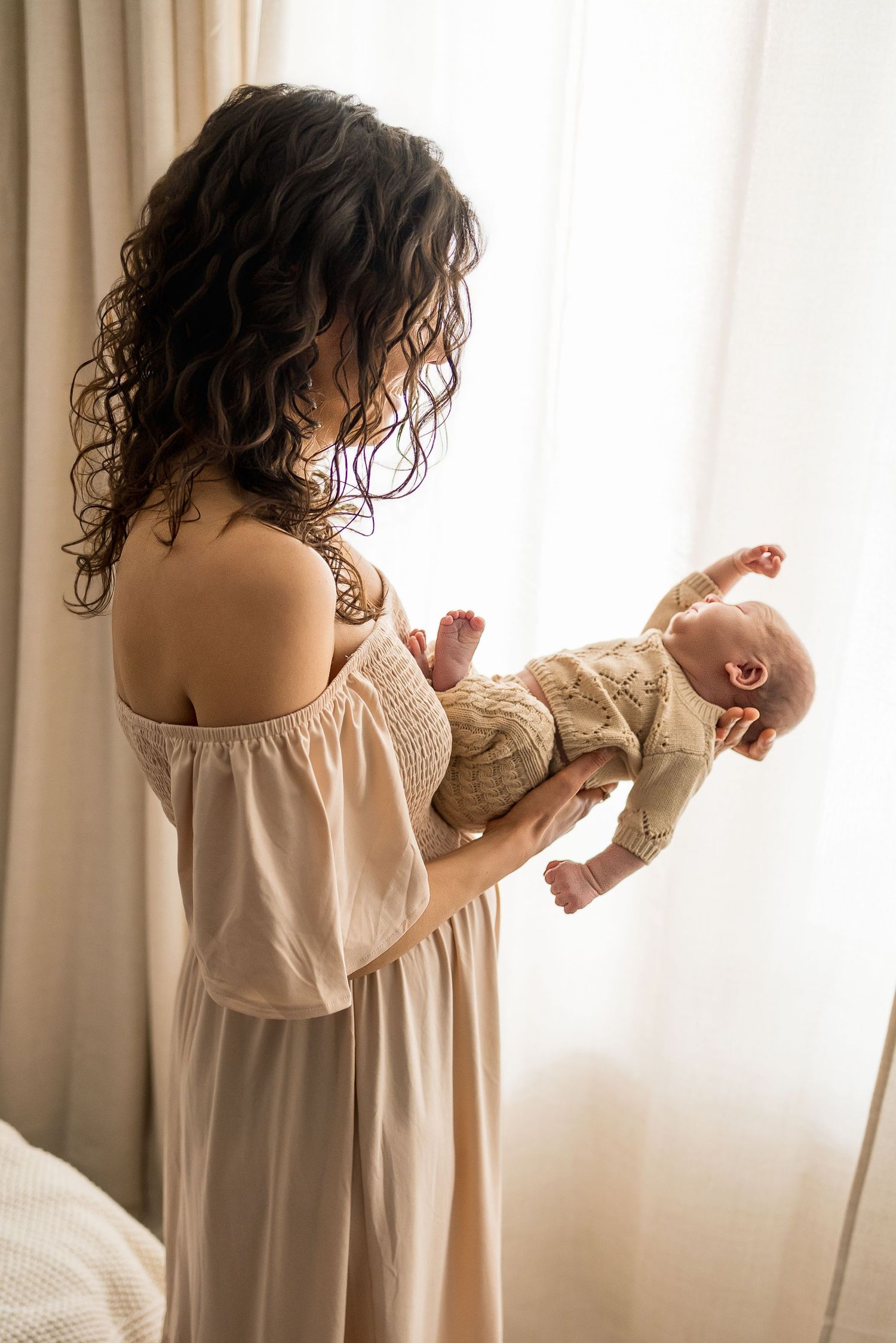 A parent in a beige off-shoulder dress holds a yawning baby wearing a knit sweater by a curtained window.