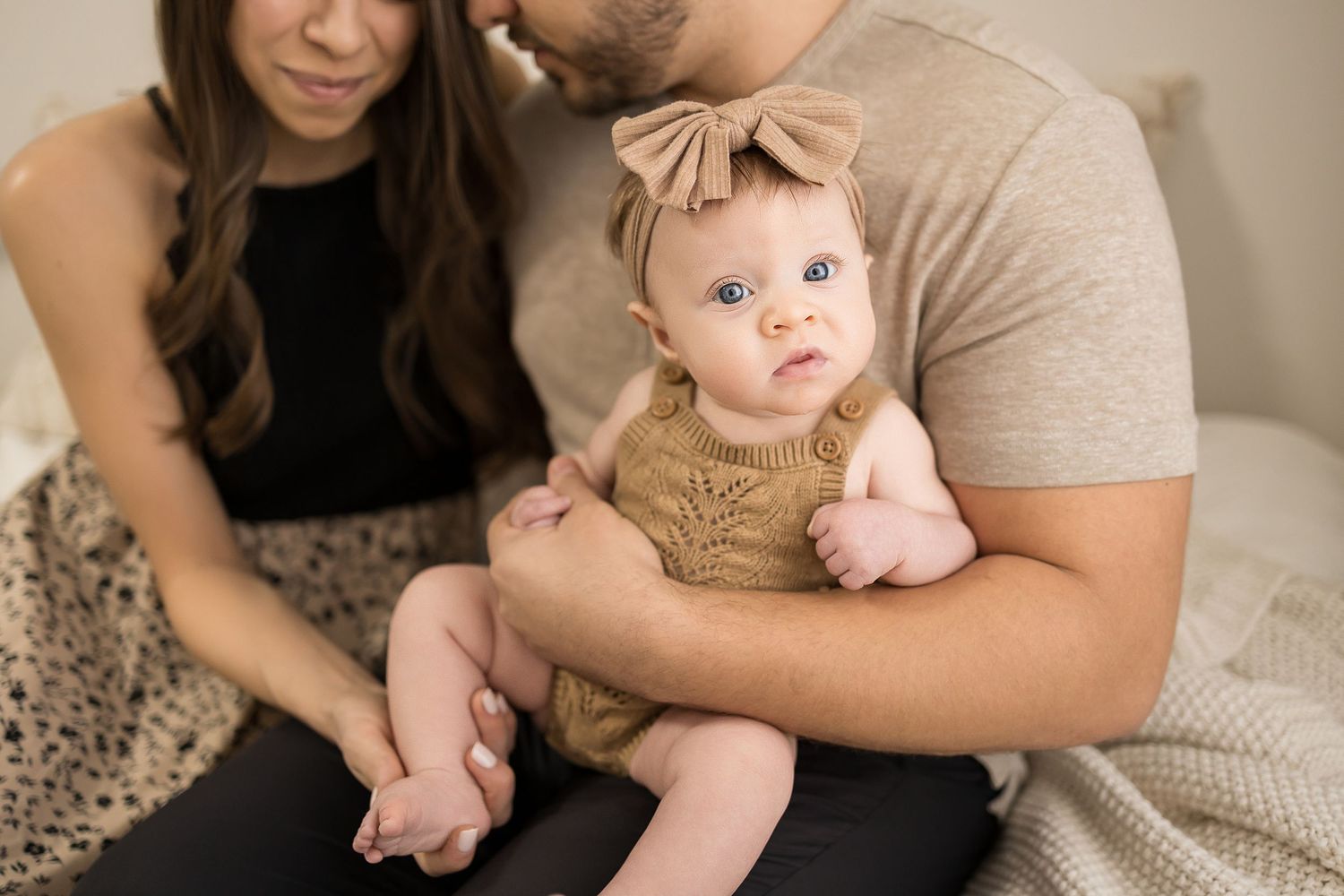 A couple lovingly holds their blue-eyed baby girl in a tan knit romper and bow headband during a family portrait session.