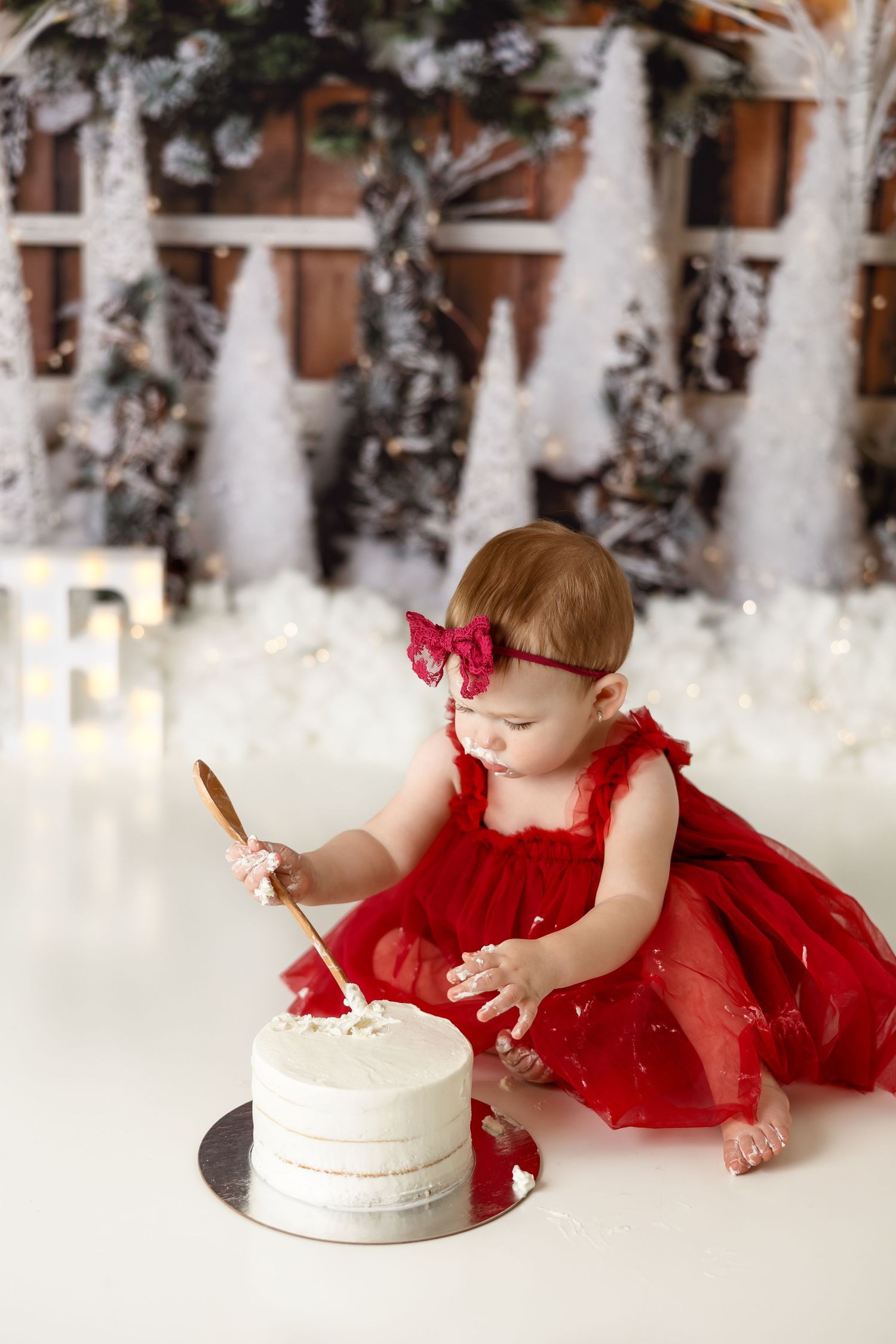 Baby in red dress and bow reaching for white cake during winter-themed first birthday photo shoot.