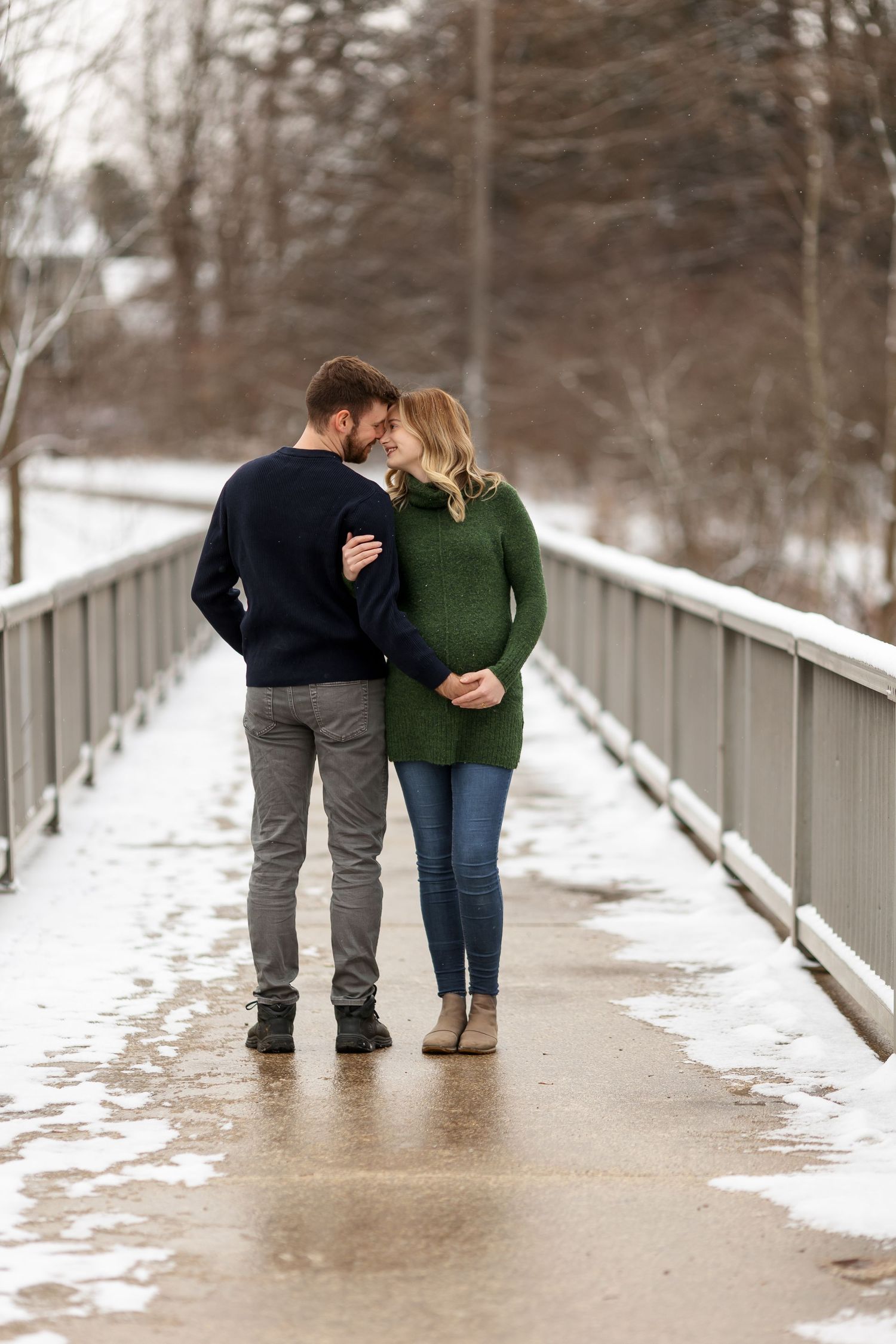 Couple standing close together on snowy bridge during winter photoshoot.