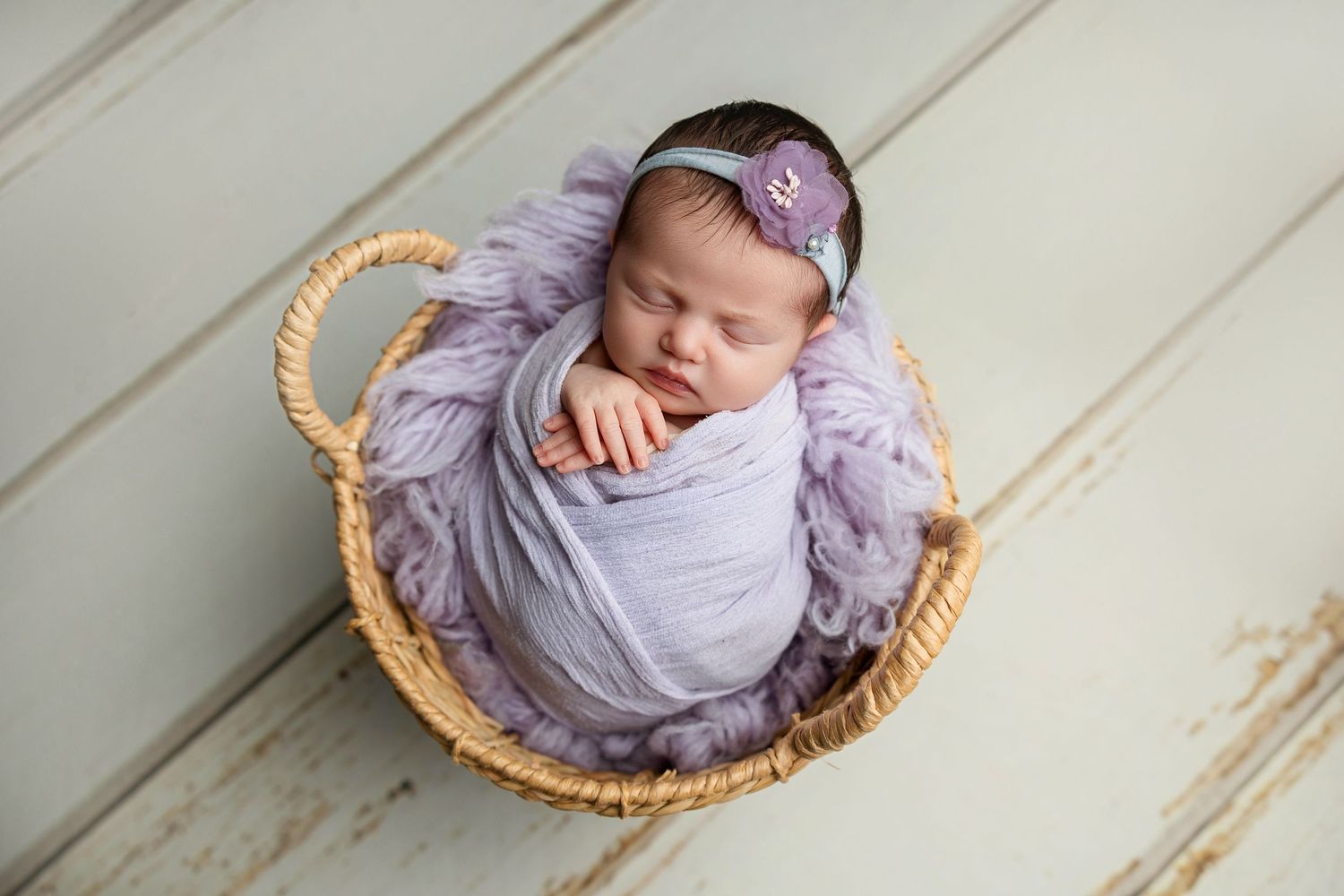 Peaceful newborn wrapped in purple swaddle sleeping in woven basket against wooden backdrop.