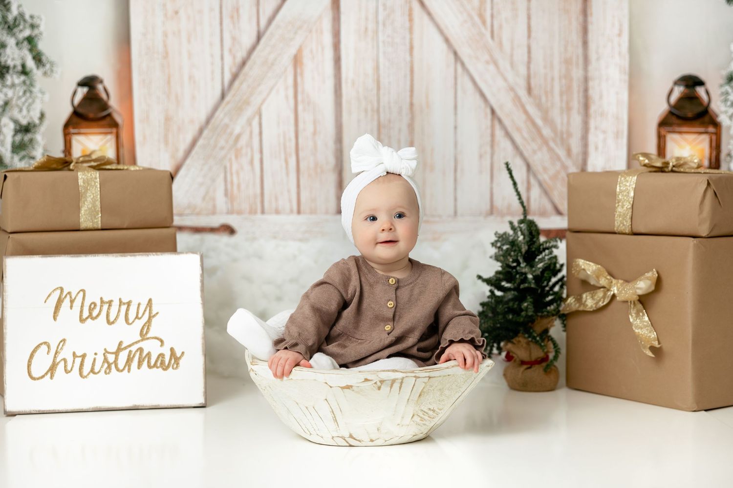 Baby in brown outfit sits in white bowl surrounded by Christmas decorations and wrapped presents.
