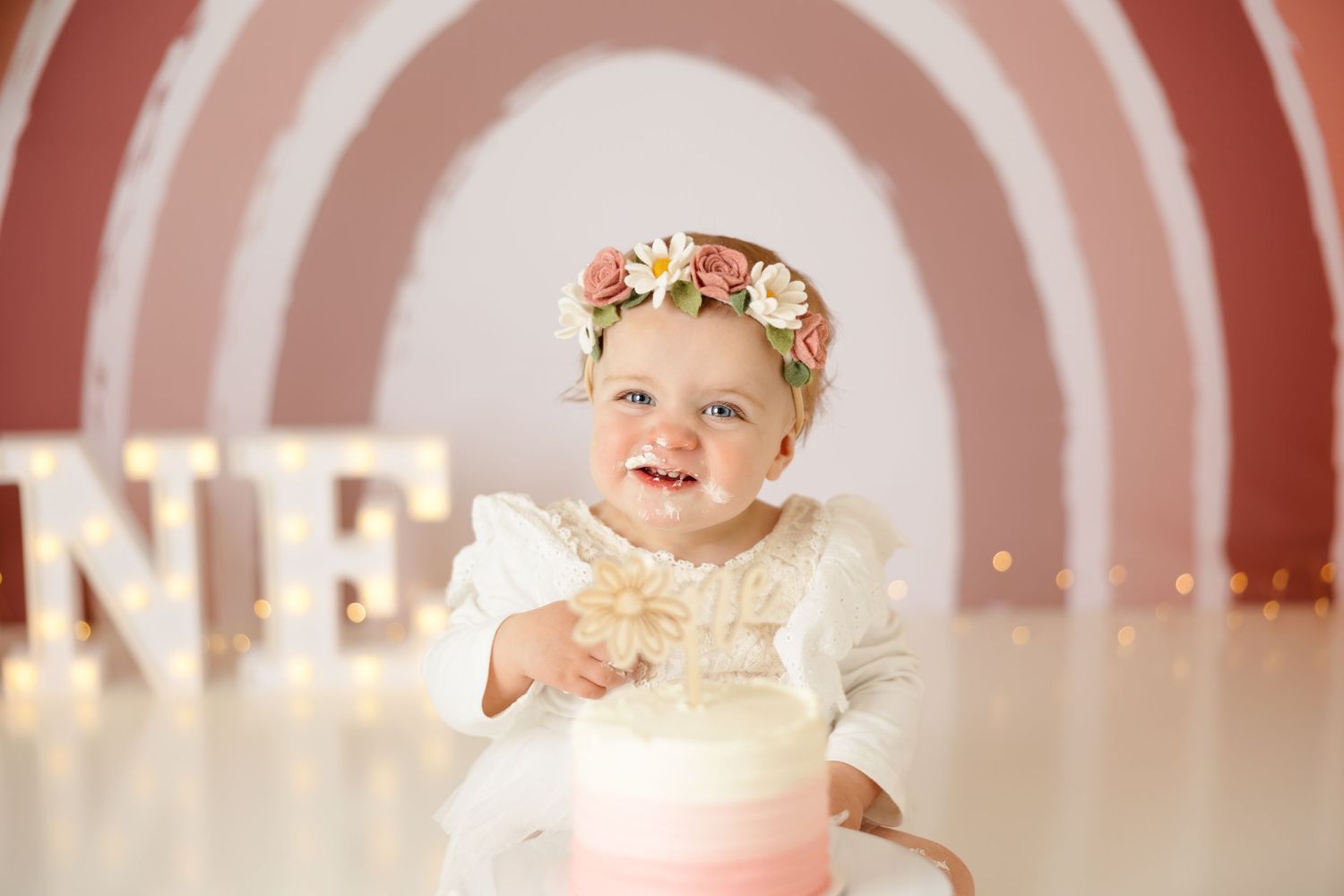 Baby's first birthday portrait with pink rainbow backdrop and elegant white dress.