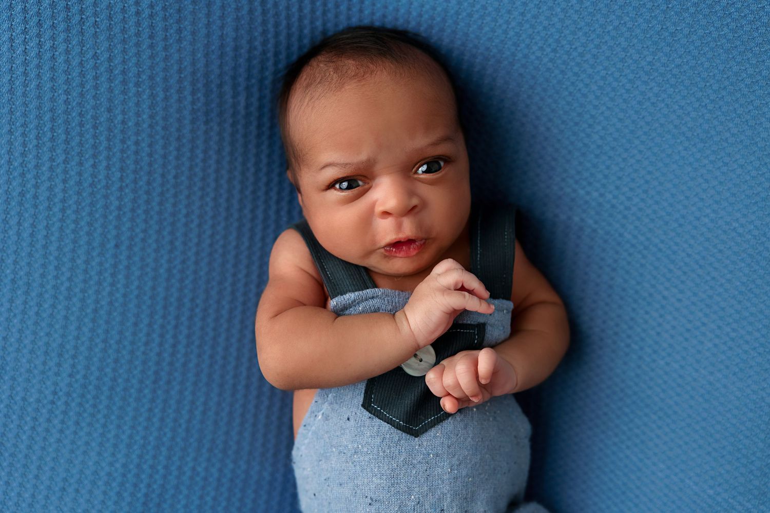 Alert baby wearing blue overalls looks at camera while resting on blue background.