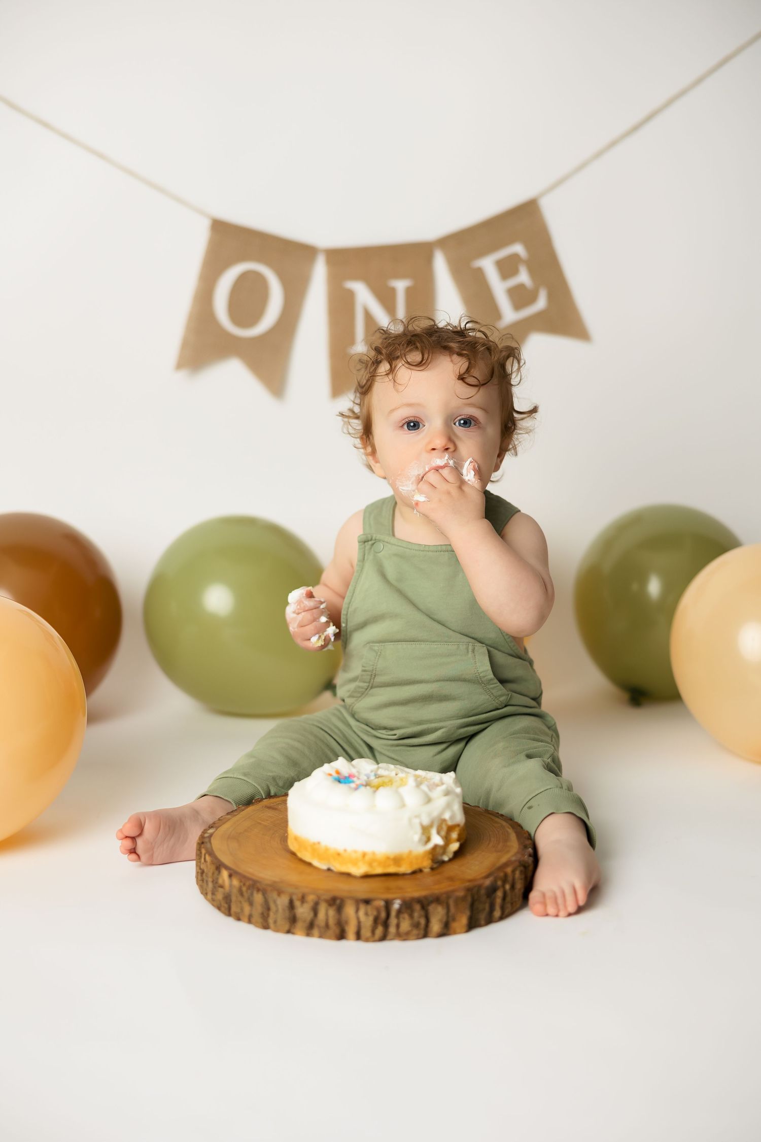 Baby in sage romper having cake smash photo session with ONE banner and balloons.
