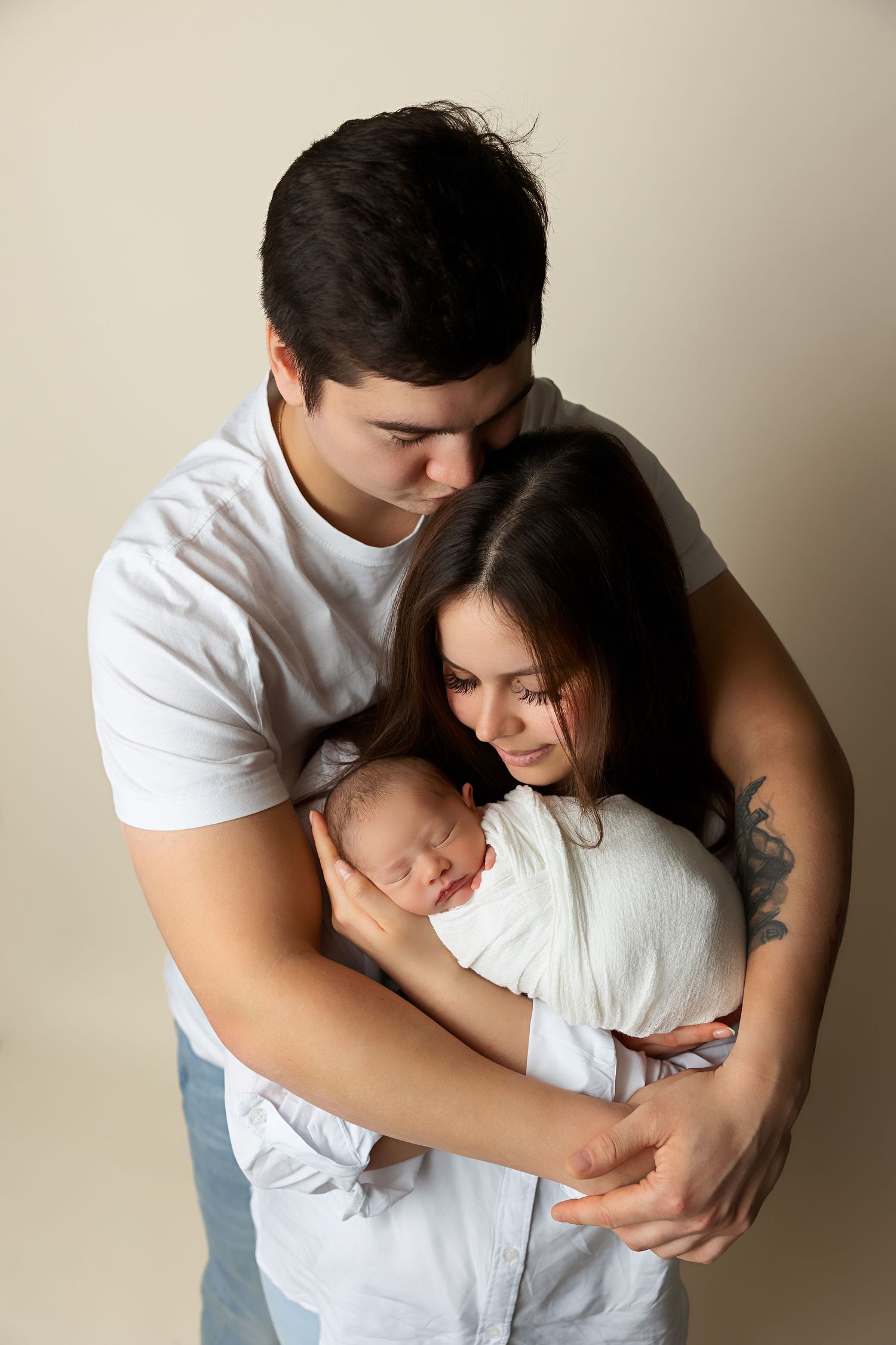 Young couple embraces their newborn baby in an intimate family portrait against neutral background.