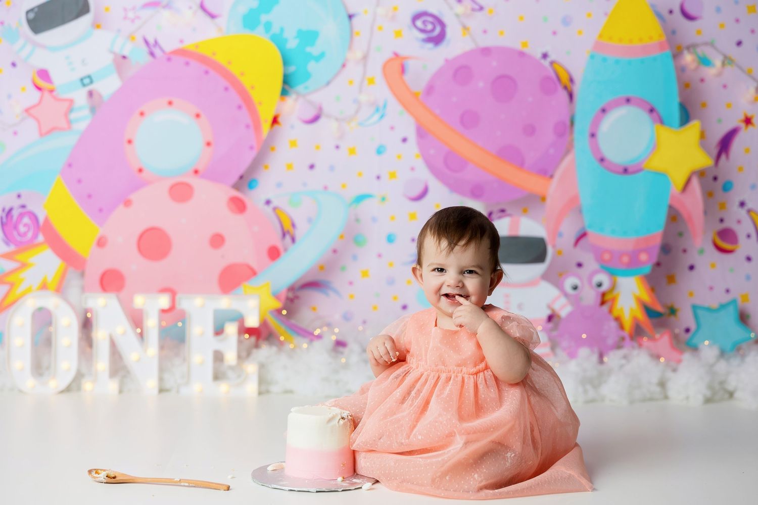 Colorful first birthday cake smash session with pink dress against white backdrop with festive decorations.