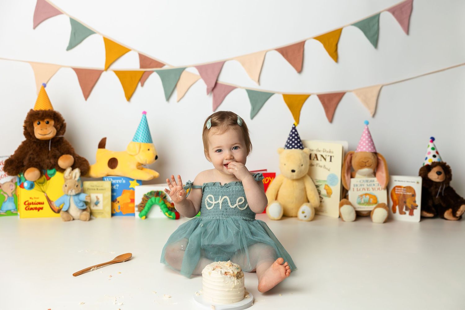 Baby girl in teal tutu dress celebrates first birthday with cake smash, surrounded by stuffed animals and colorful books.