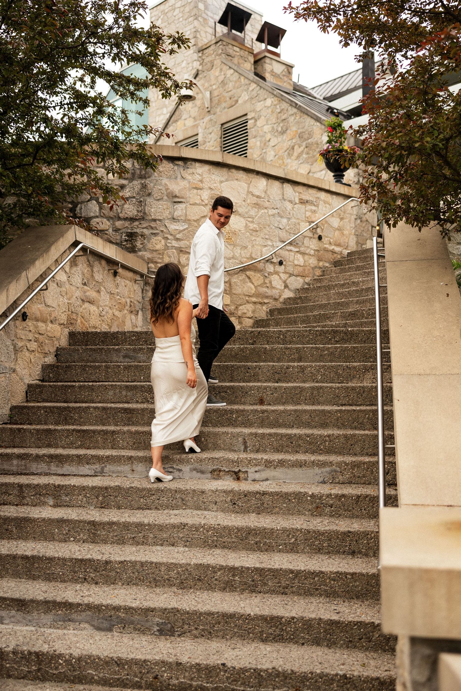 Couple walking up stone stairs, woman in white backless dress and man in white shirt looking back at her.