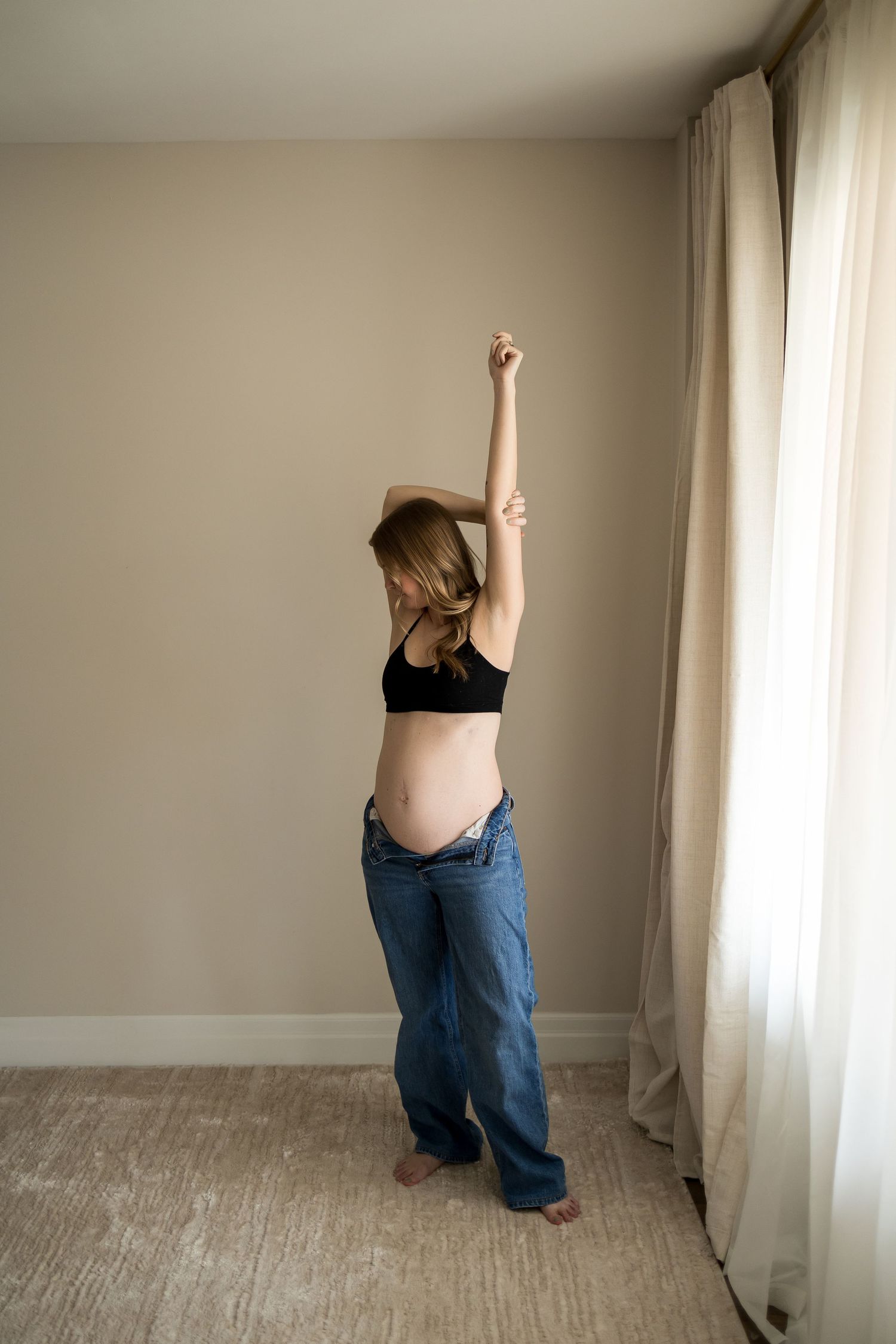 Pregnant woman in black crop top and loose jeans stretching arms overhead near a curtained window.