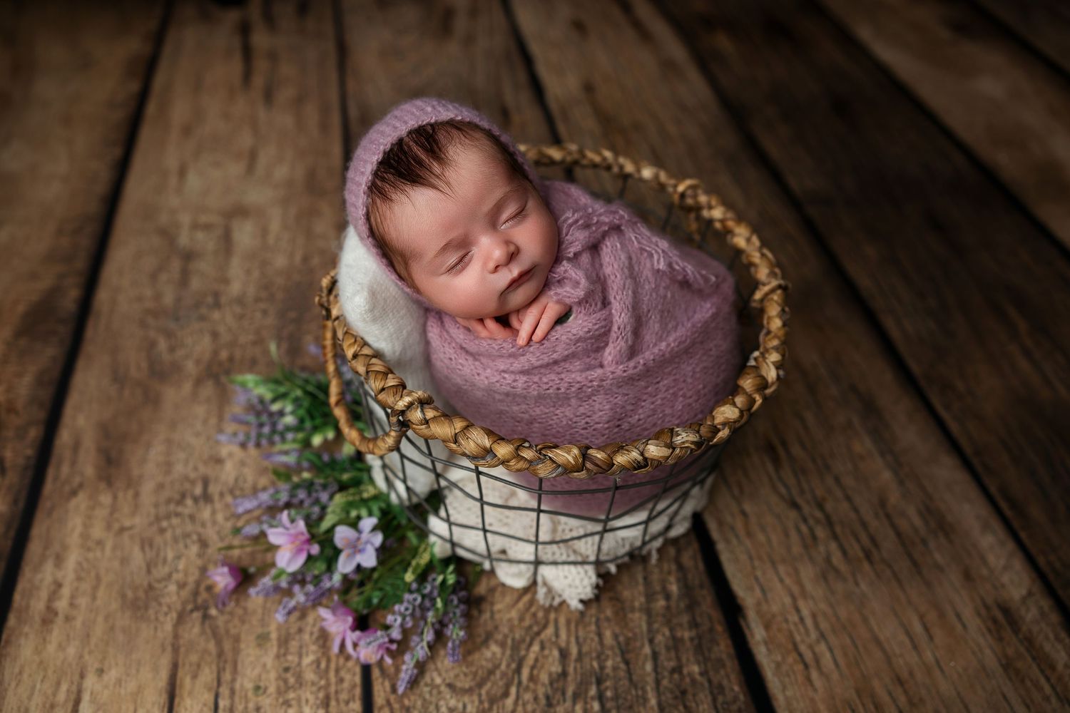 Peaceful newborn sleeping in rustic basket with purple wildflowers.