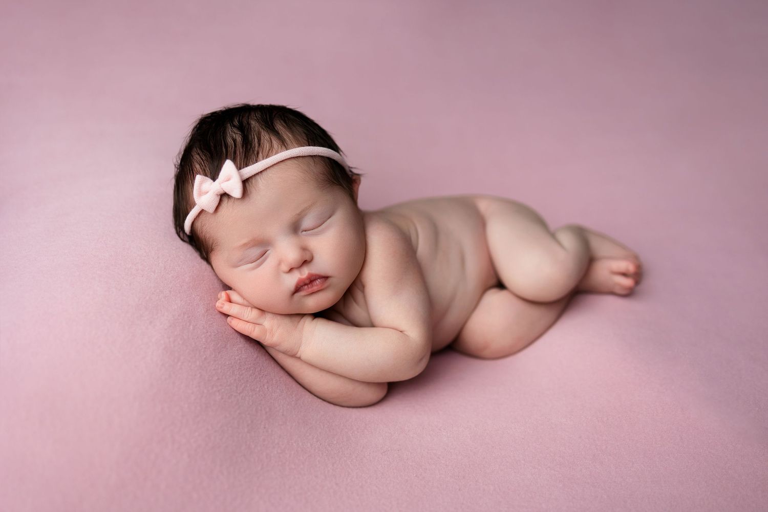 A sleeping newborn with white bow headband poses on pink backdrop.