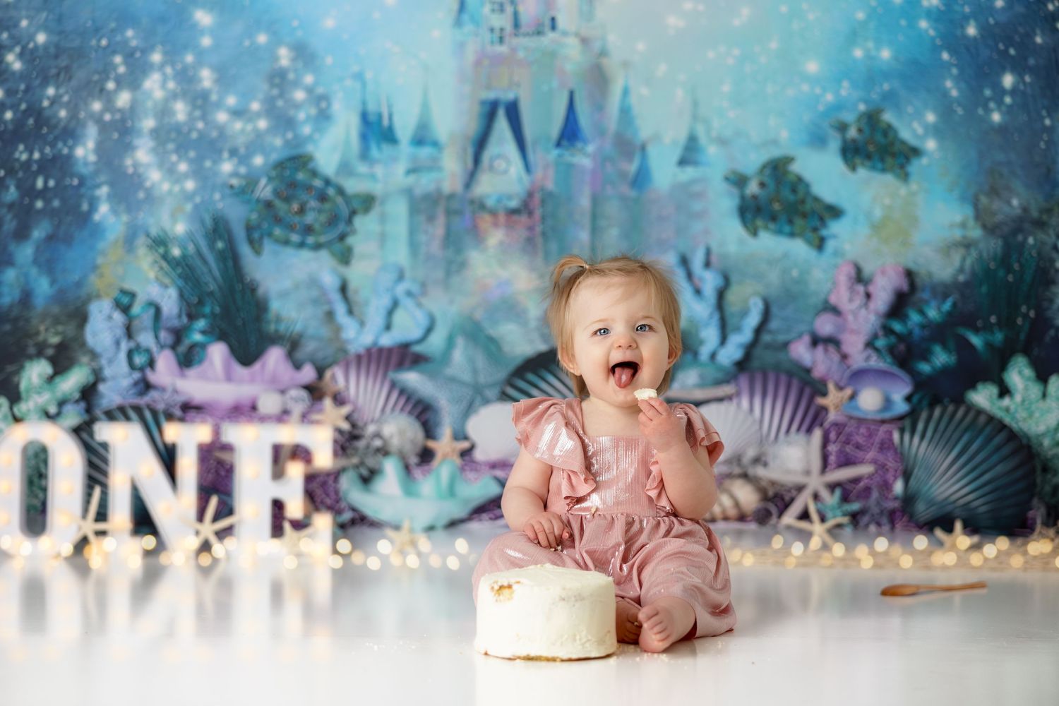 Baby in dusty rose outfit celebrates with cake against magical underwater themed backdrop.