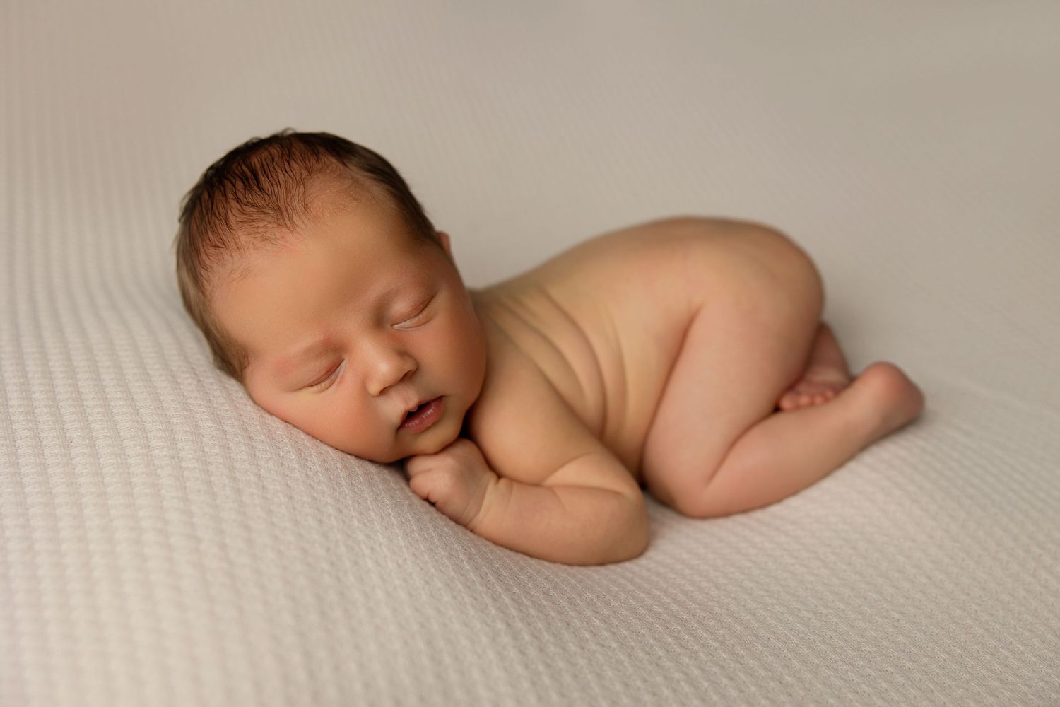 Sleeping newborn curled up on side against cream textured backdrop.