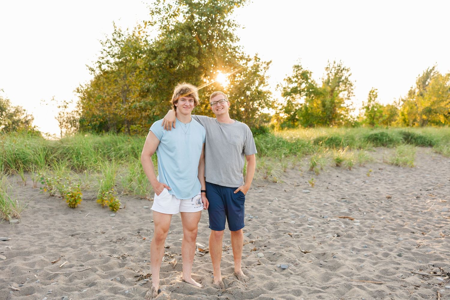 Two people standing together on a sandy beach during golden hour sunset with trees in the background.