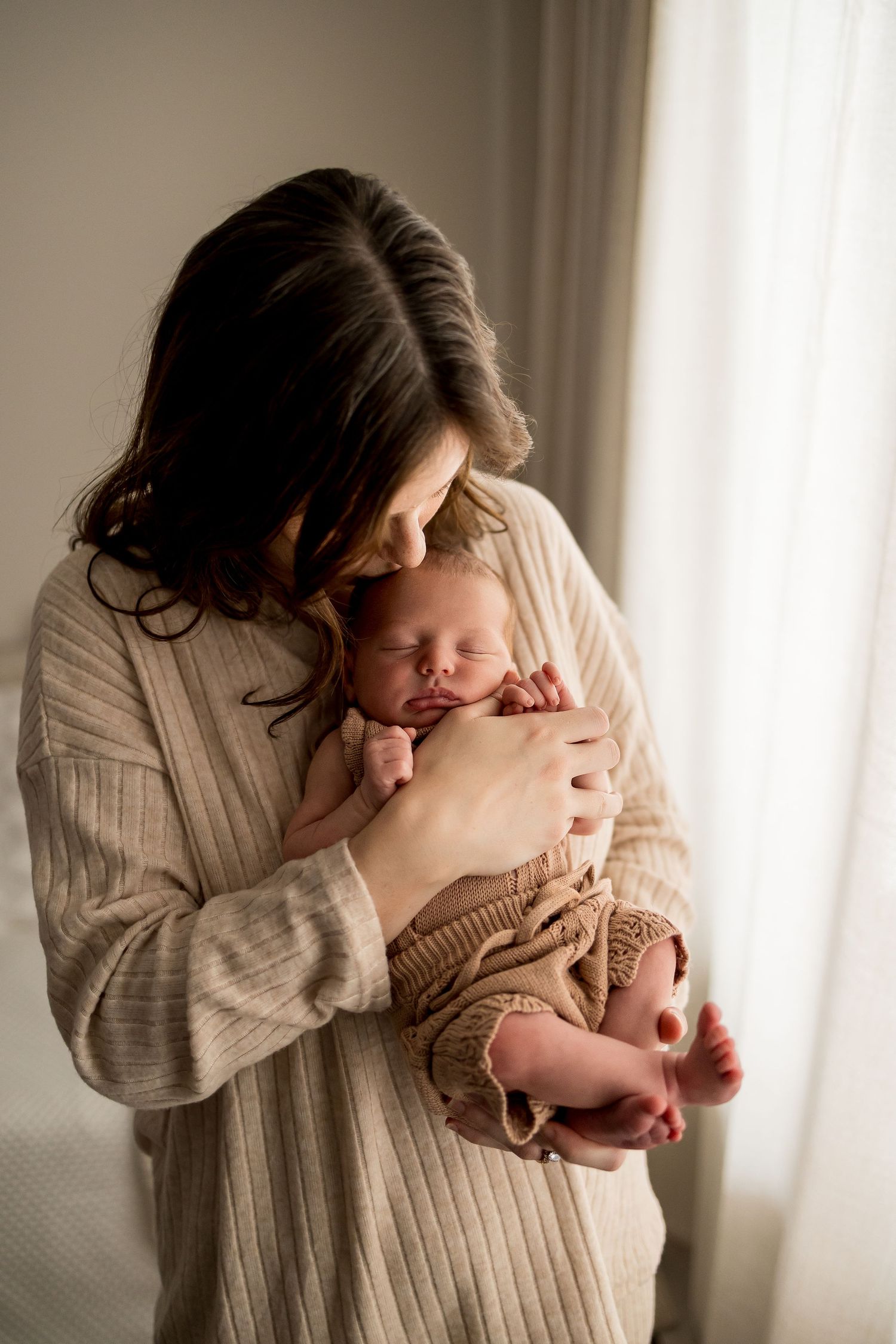 Tender moment by window light as parent in beige sweater cradles sleeping newborn baby in warm embrace.