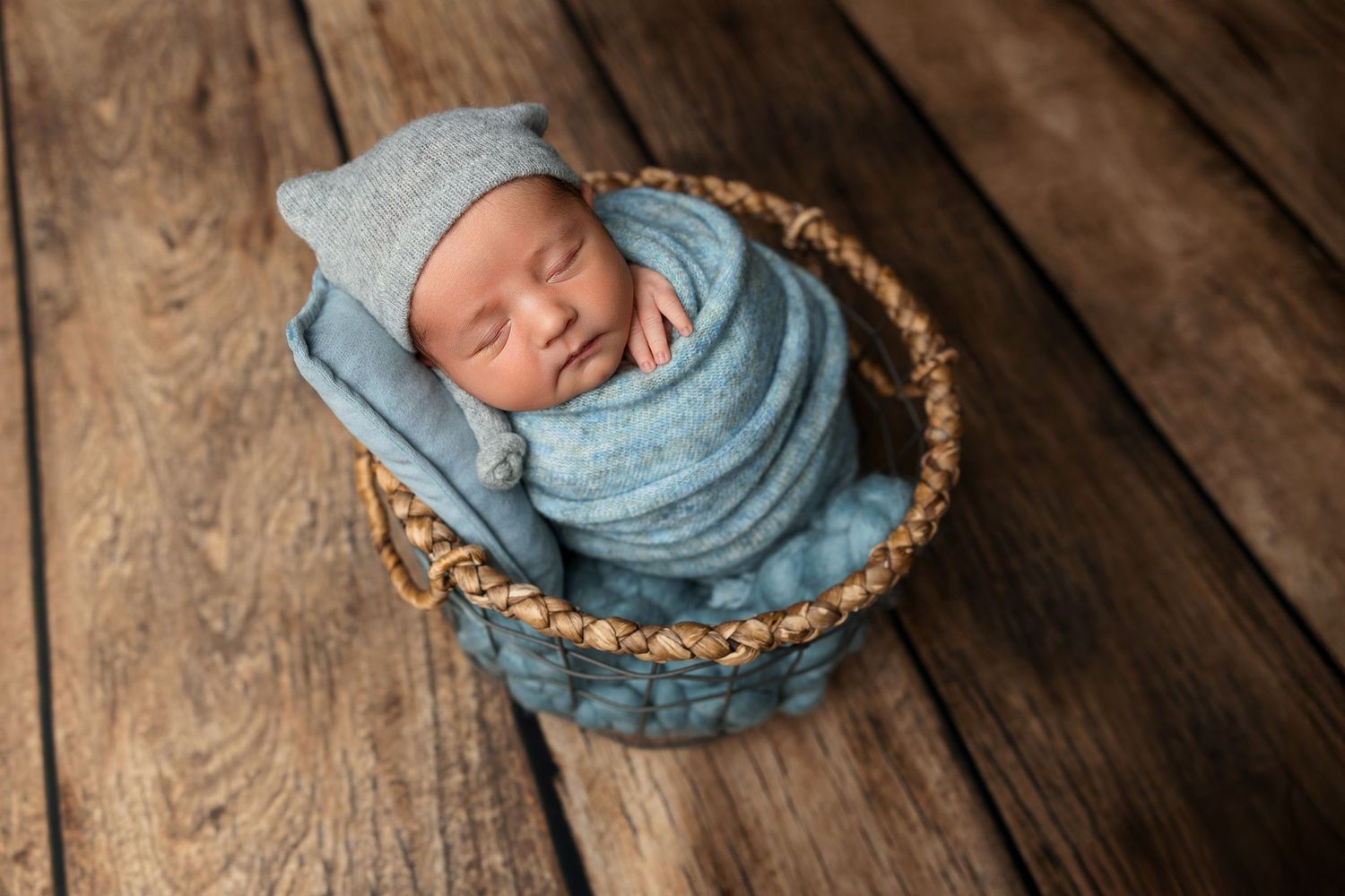 Sleeping newborn wrapped in blue swaddle resting in woven basket on wooden floor.