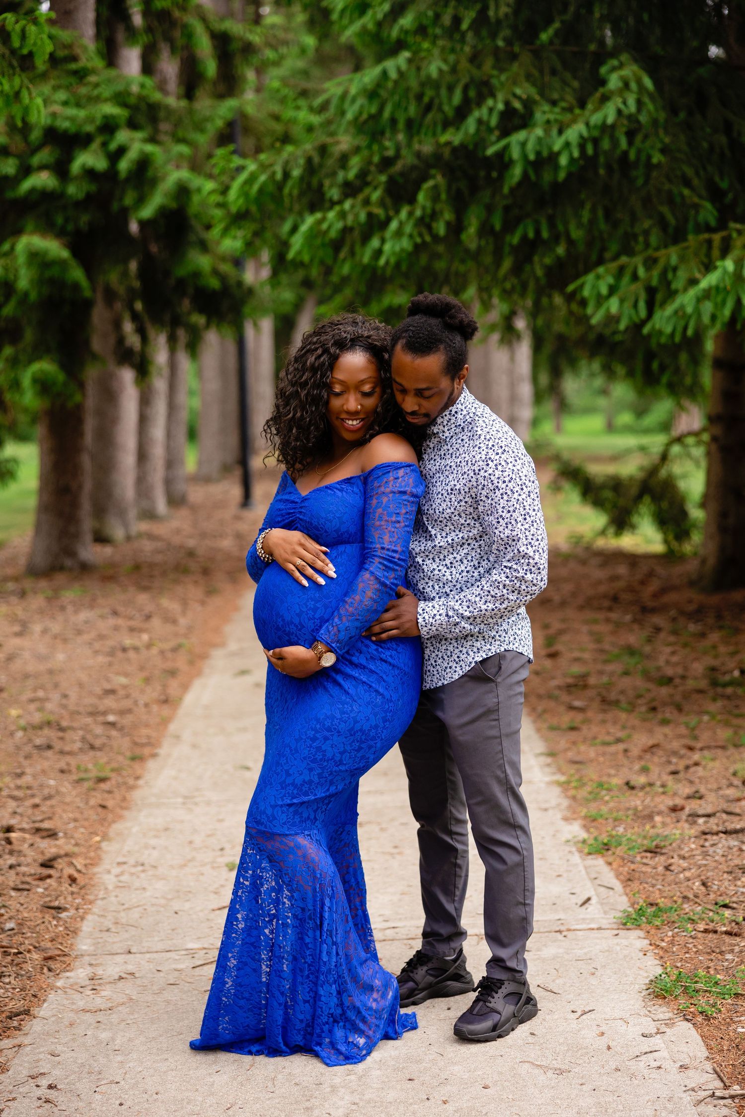 Maternity photos of couple in blue dress and patterned shirt walking through pine forest.