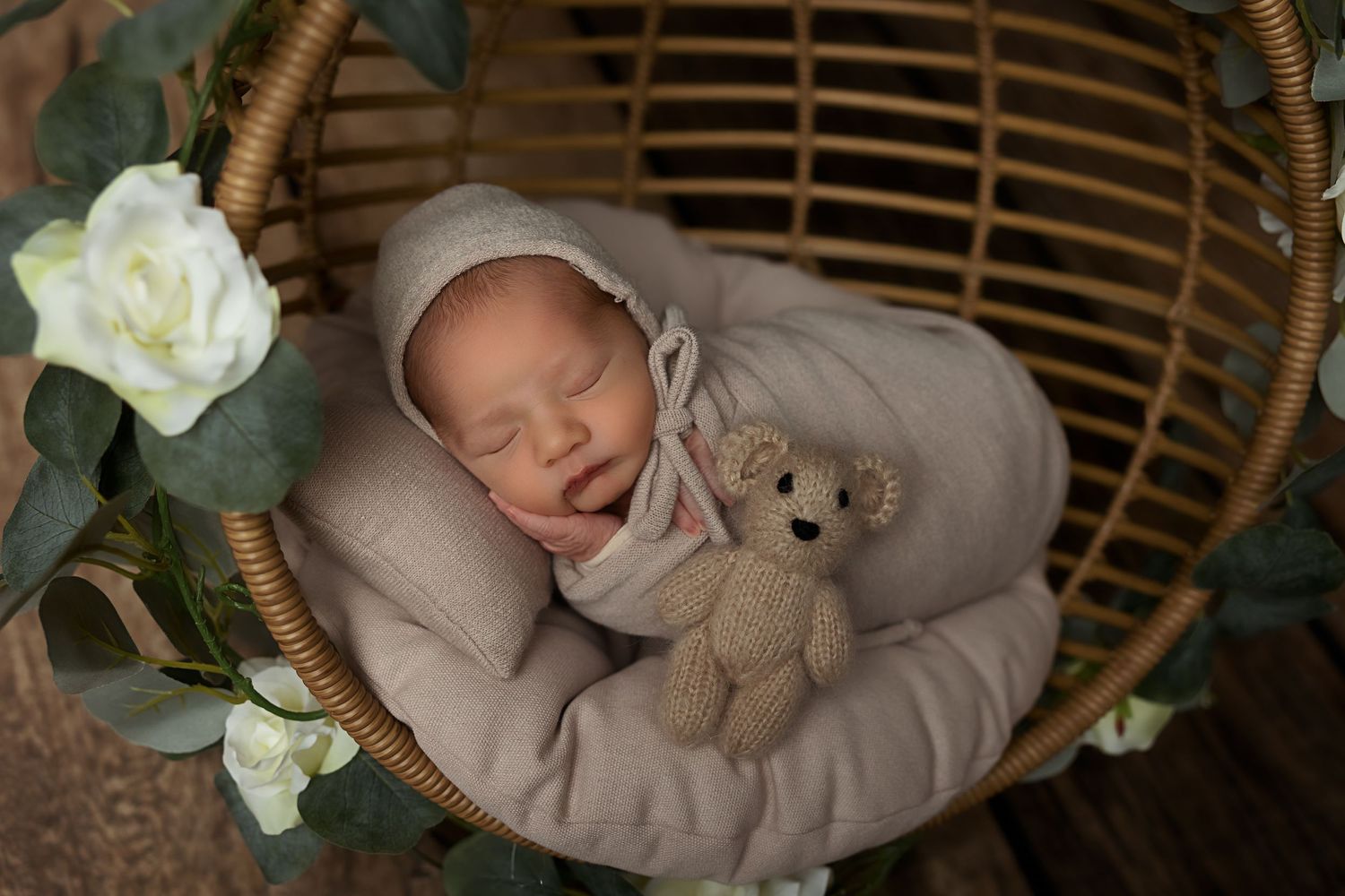 Newborn sleeping in rattan chair with teddy bear and white roses.