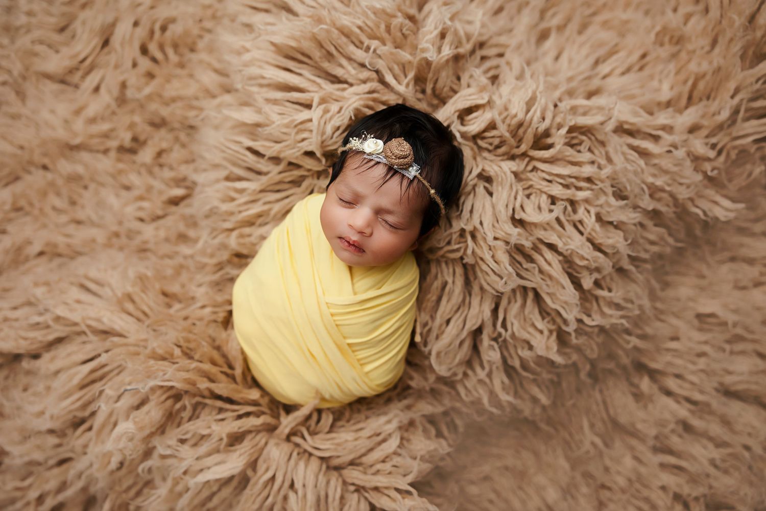 Newborn wrapped in yellow swaddle sleeping on textured beige fur backdrop.