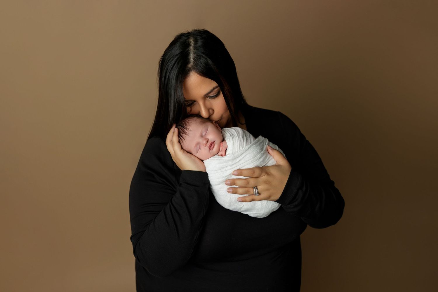 Mother in black sweater holding swaddled newborn against brown backdrop.