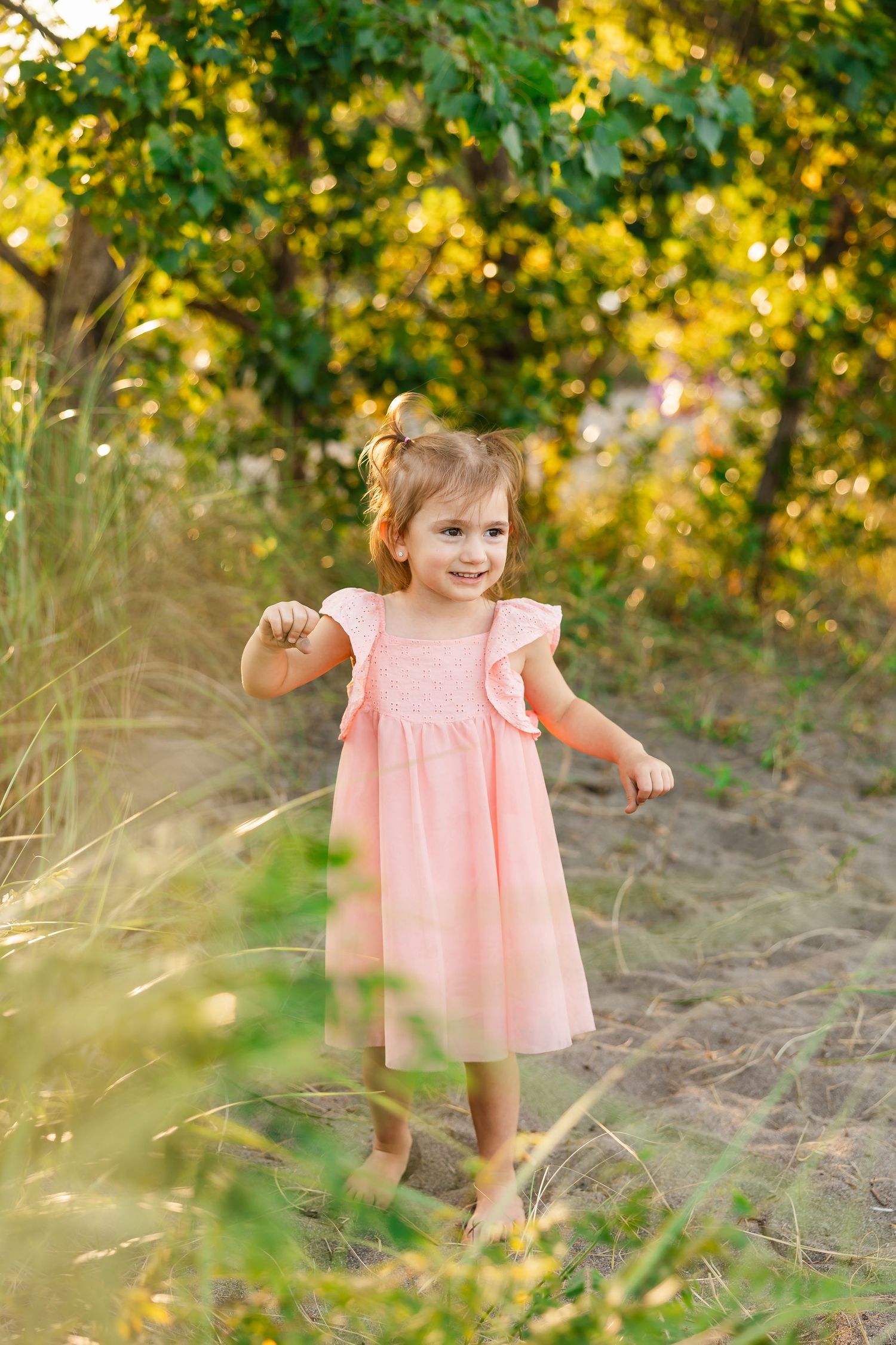A young child in a pink dress plays outdoors among trees and tall grass during golden hour lighting.