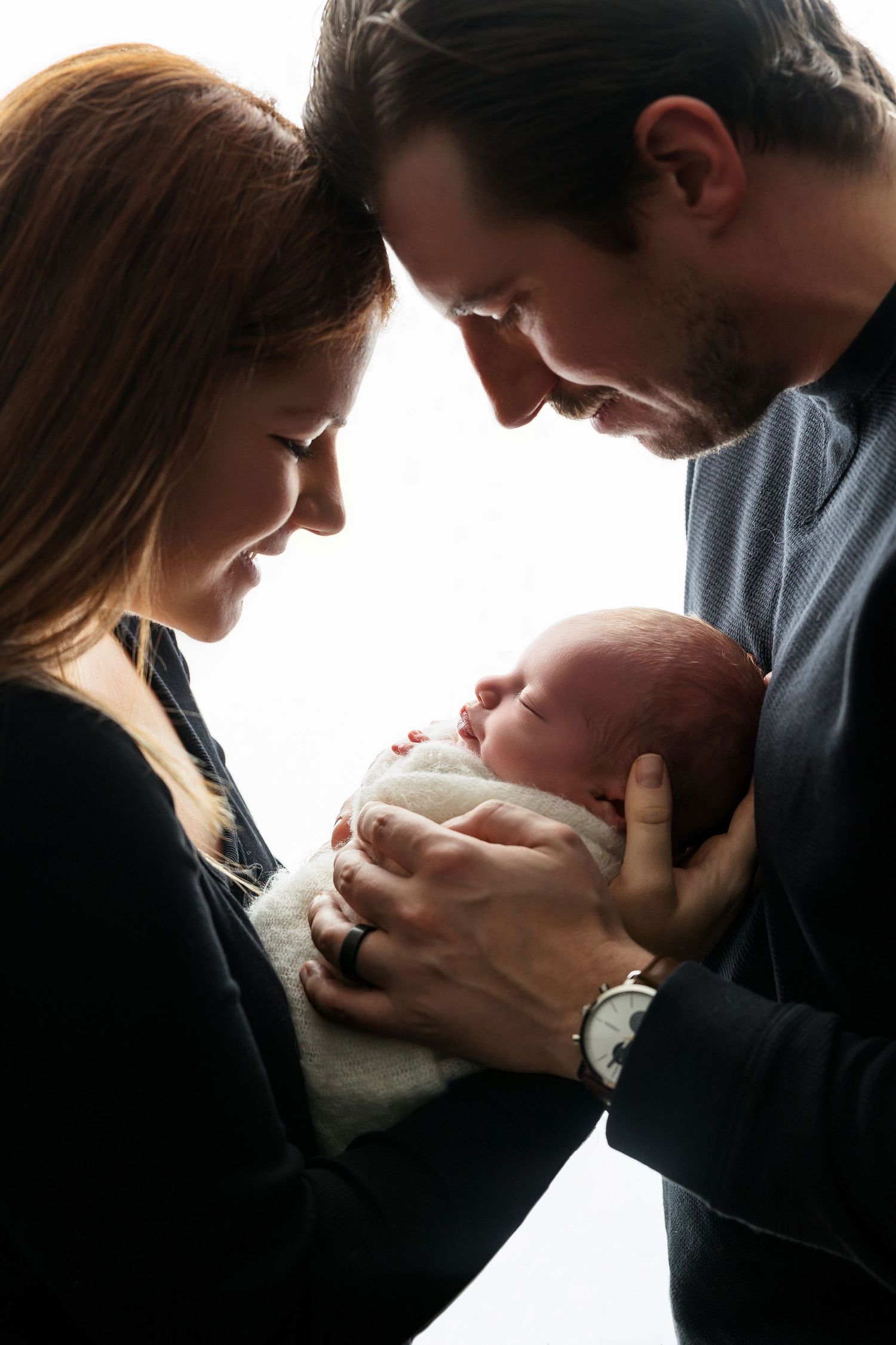 Intimate backlit portrait of parents gazing at swaddled newborn.