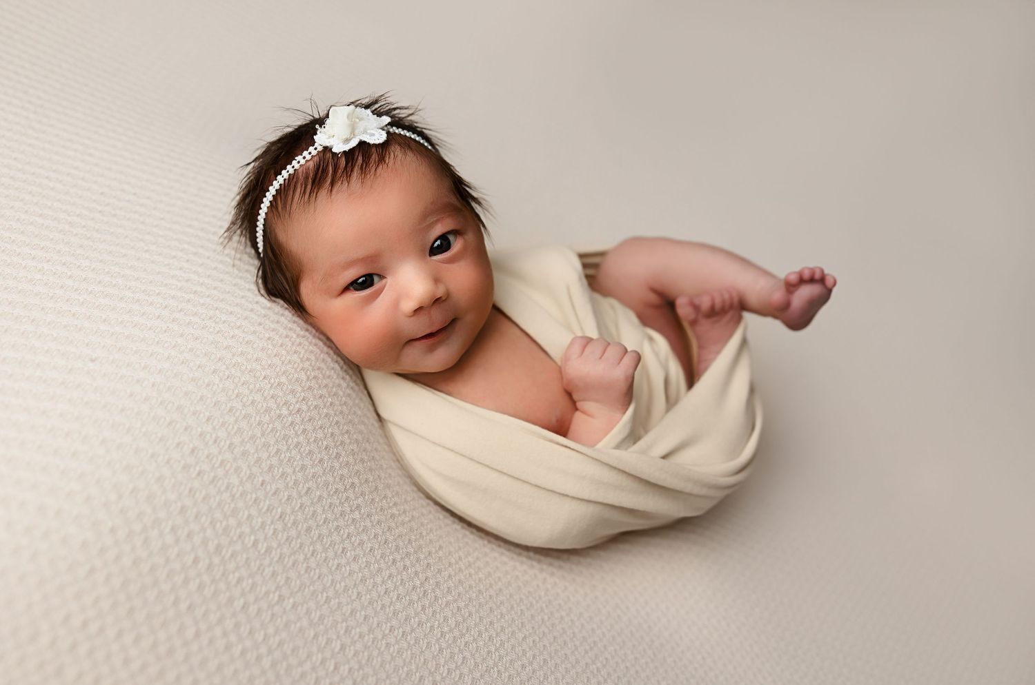 Newborn wrapped in cream fabric lying on textured white backdrop with delicate flower headband.