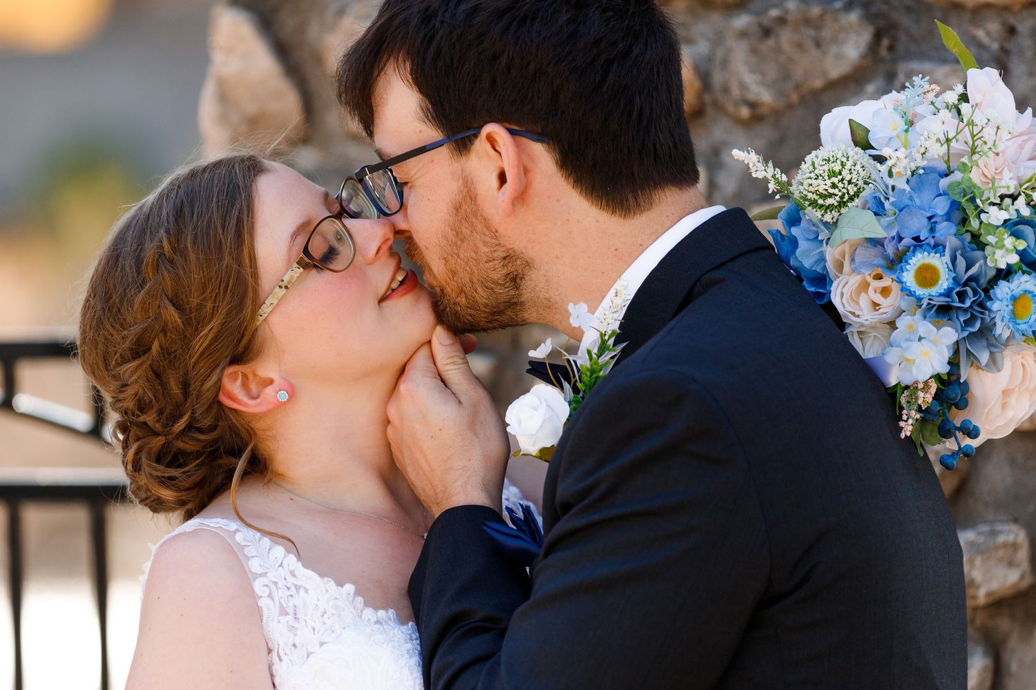 Tender wedding moment captured against stone wall with blue floral bouquet.