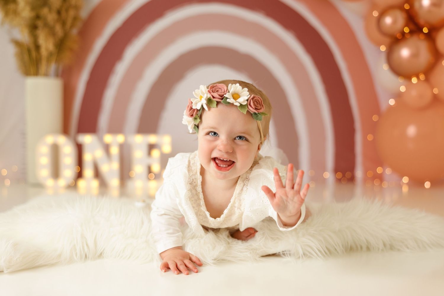 Baby waving at camera while lying on white fur with pink rainbow backdrop.