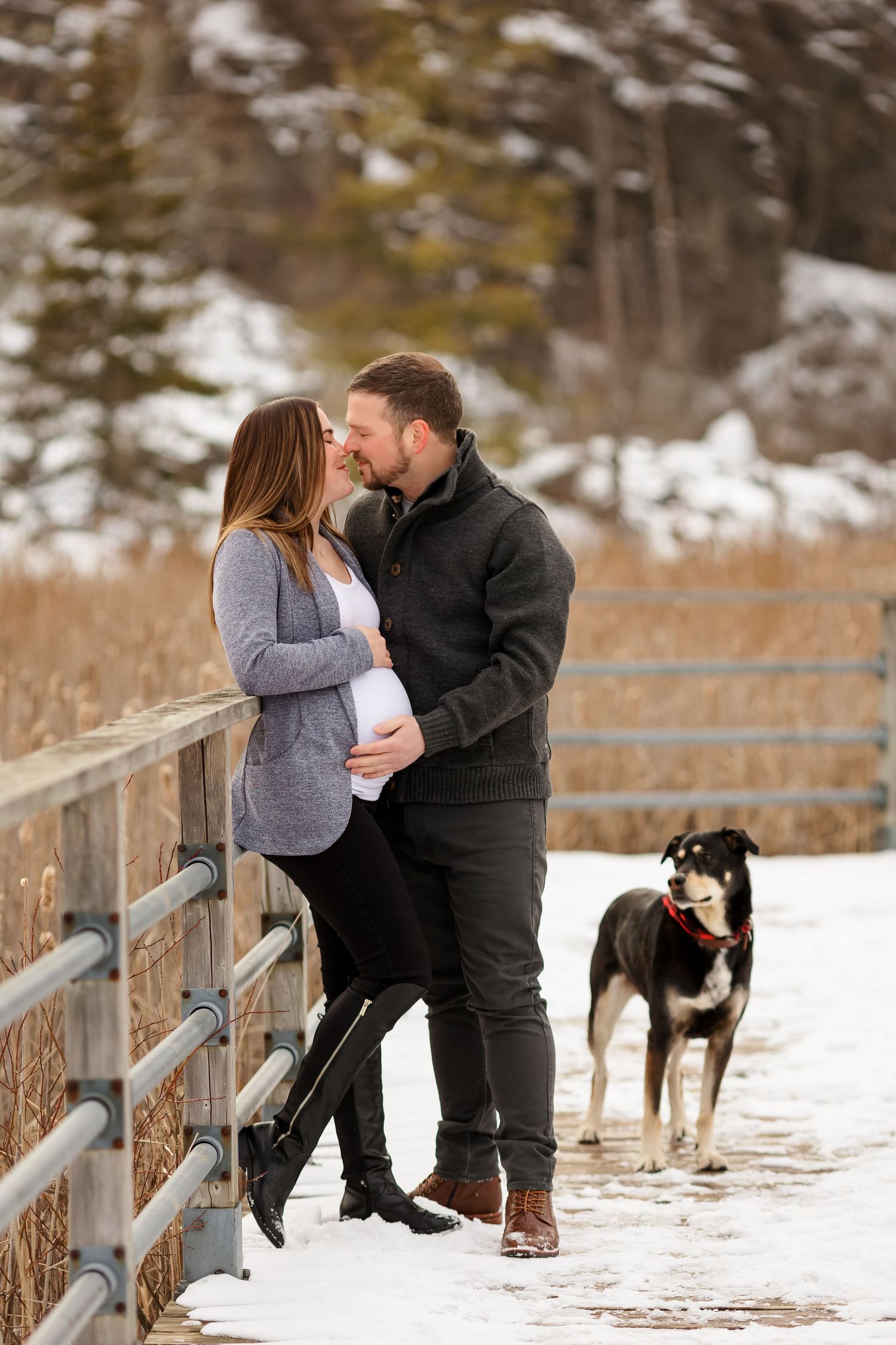 Romantic maternity photo on wooden bridge with dog watching in snowy background.