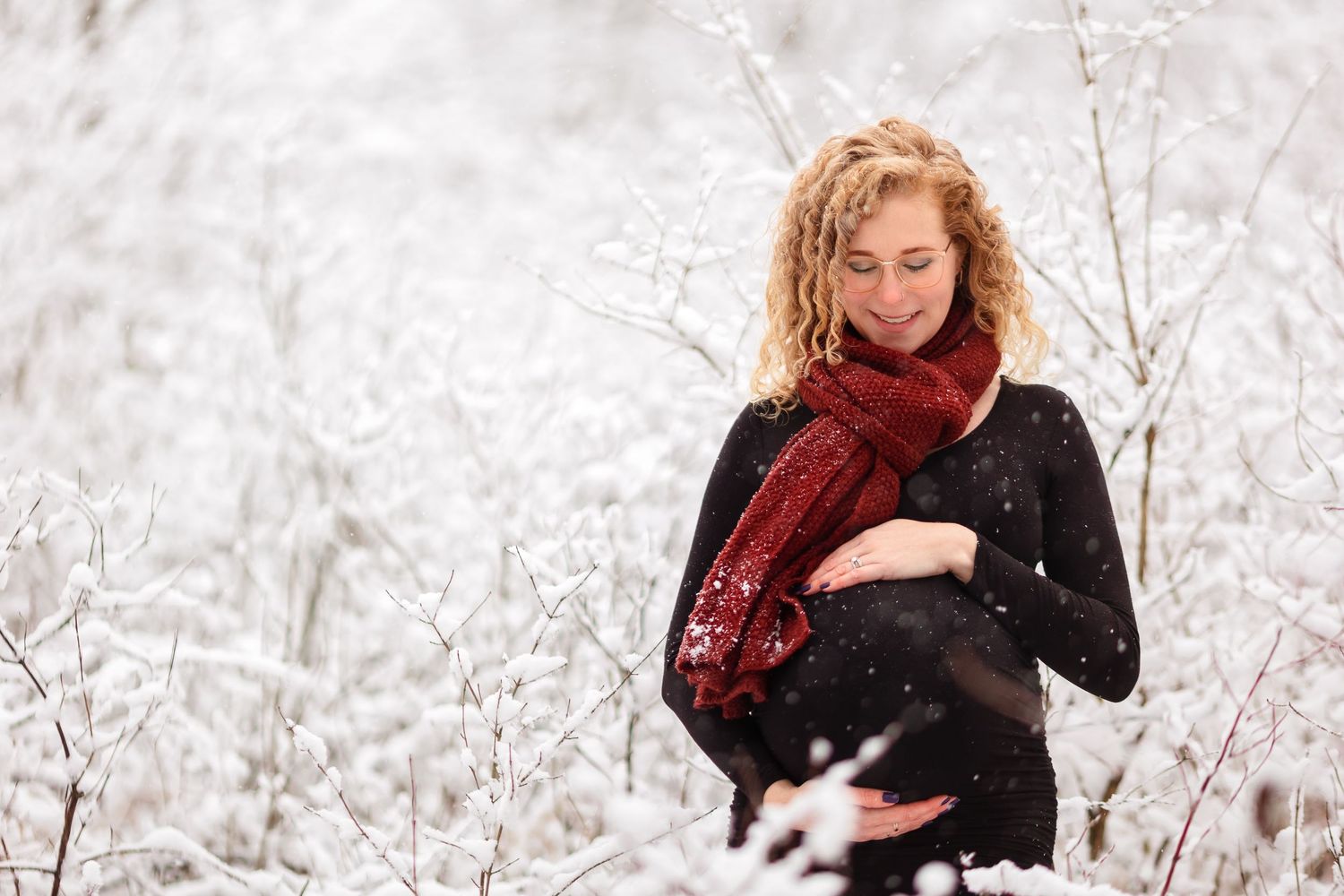 Pregnant woman in black coat and red scarf poses against snowy winter backdrop.