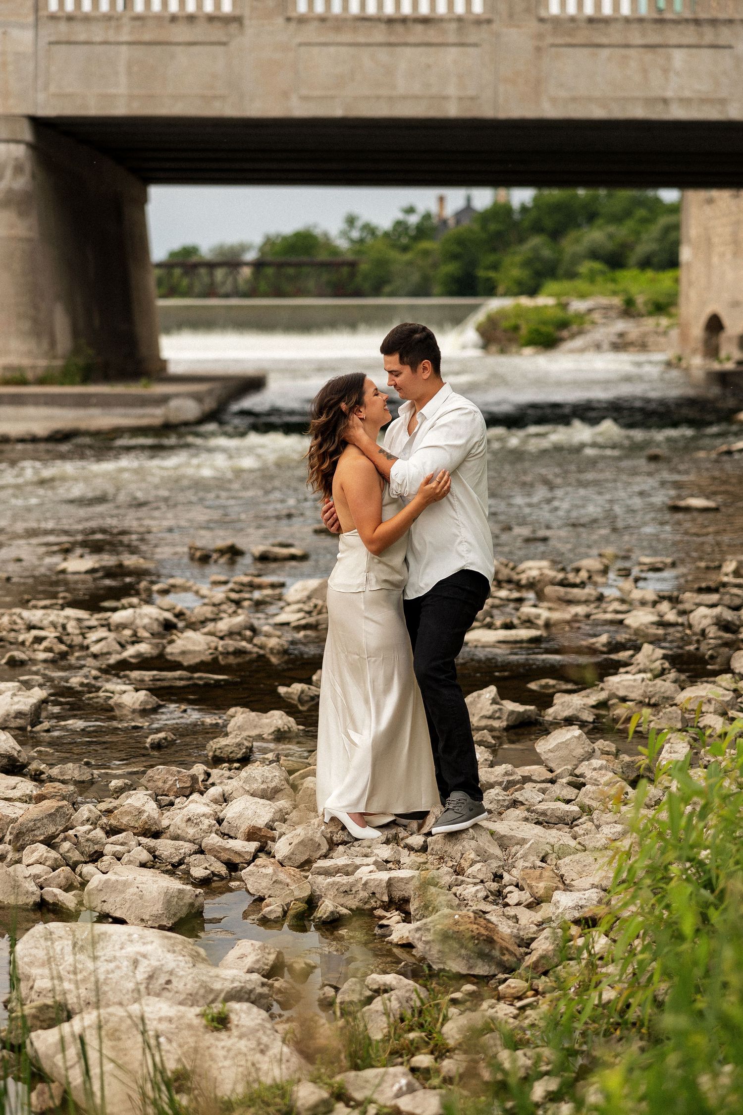 Couple embracing romantically on rocky riverbank near a bridge, woman in white dress, man in white shirt and black pants.