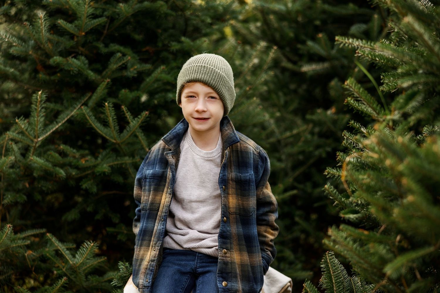 Young person wearing a beanie and plaid shirt poses in front of evergreen trees outdoors.