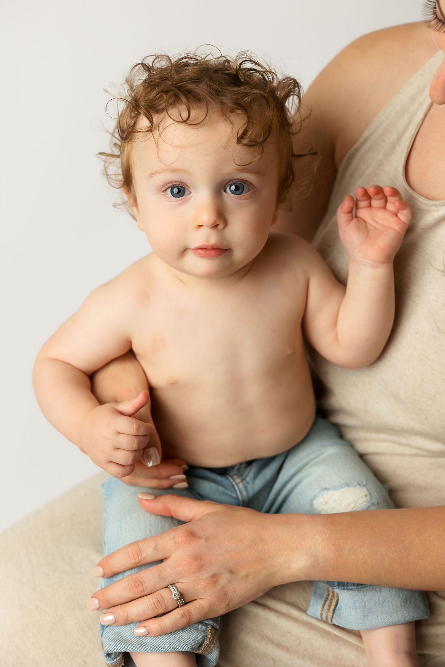 Close up portrait of curly-haired toddler in denim shorts held by adult in beige tank top.