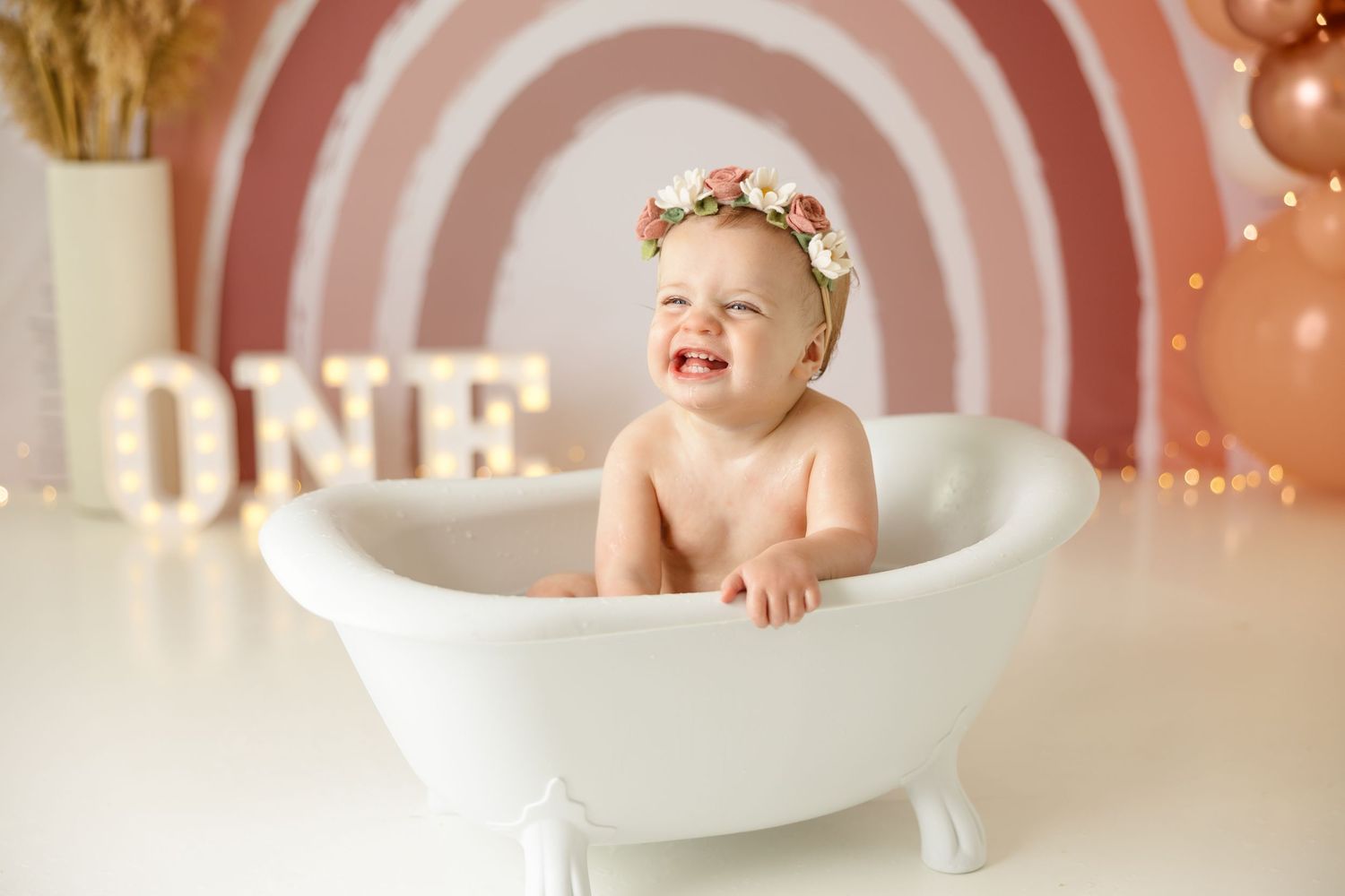 Sweet first birthday milestone photo in white clawfoot tub against rainbow backdrop.