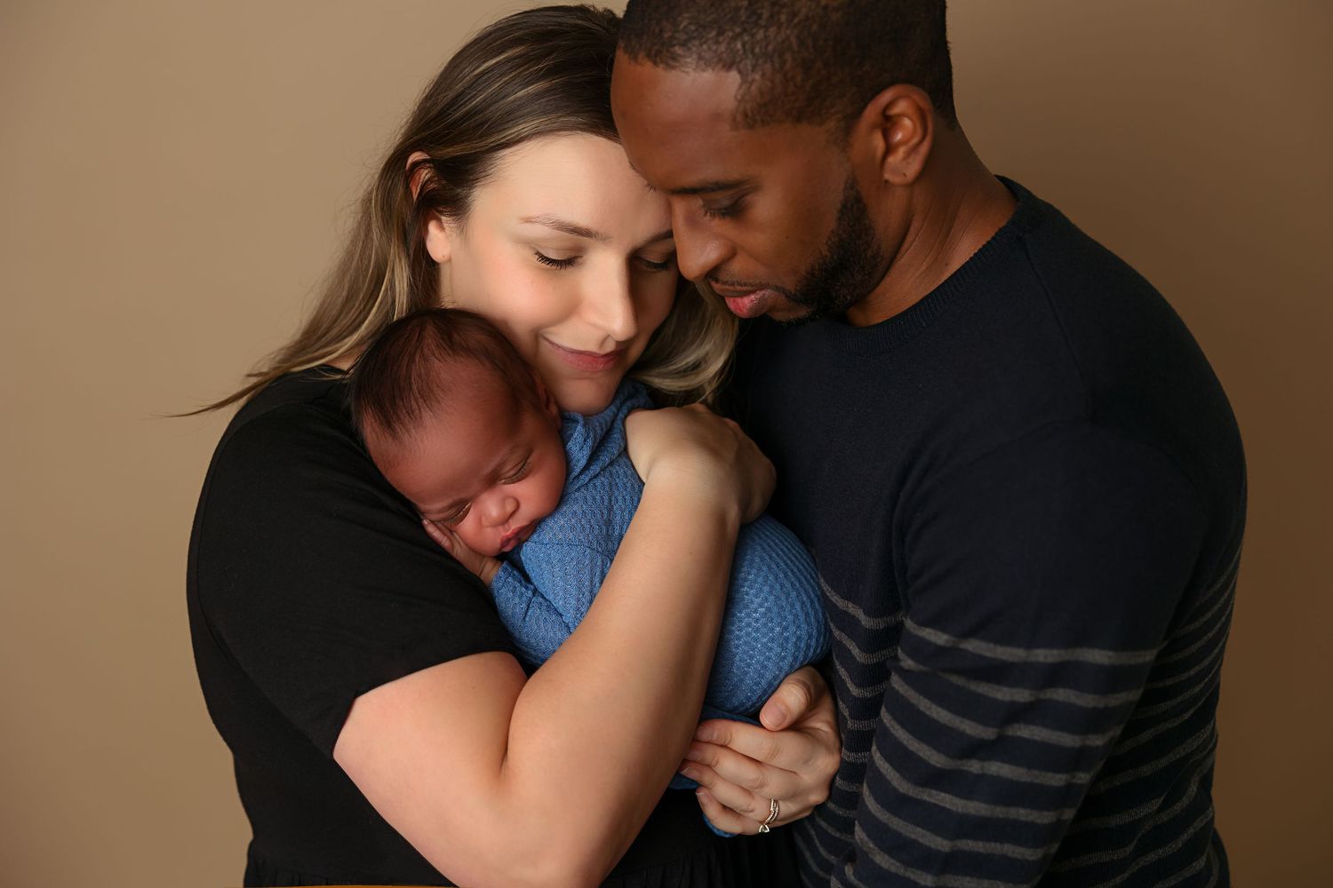 Intimate family portrait with parents embracing newborn in blue outfit against brown backdrop.