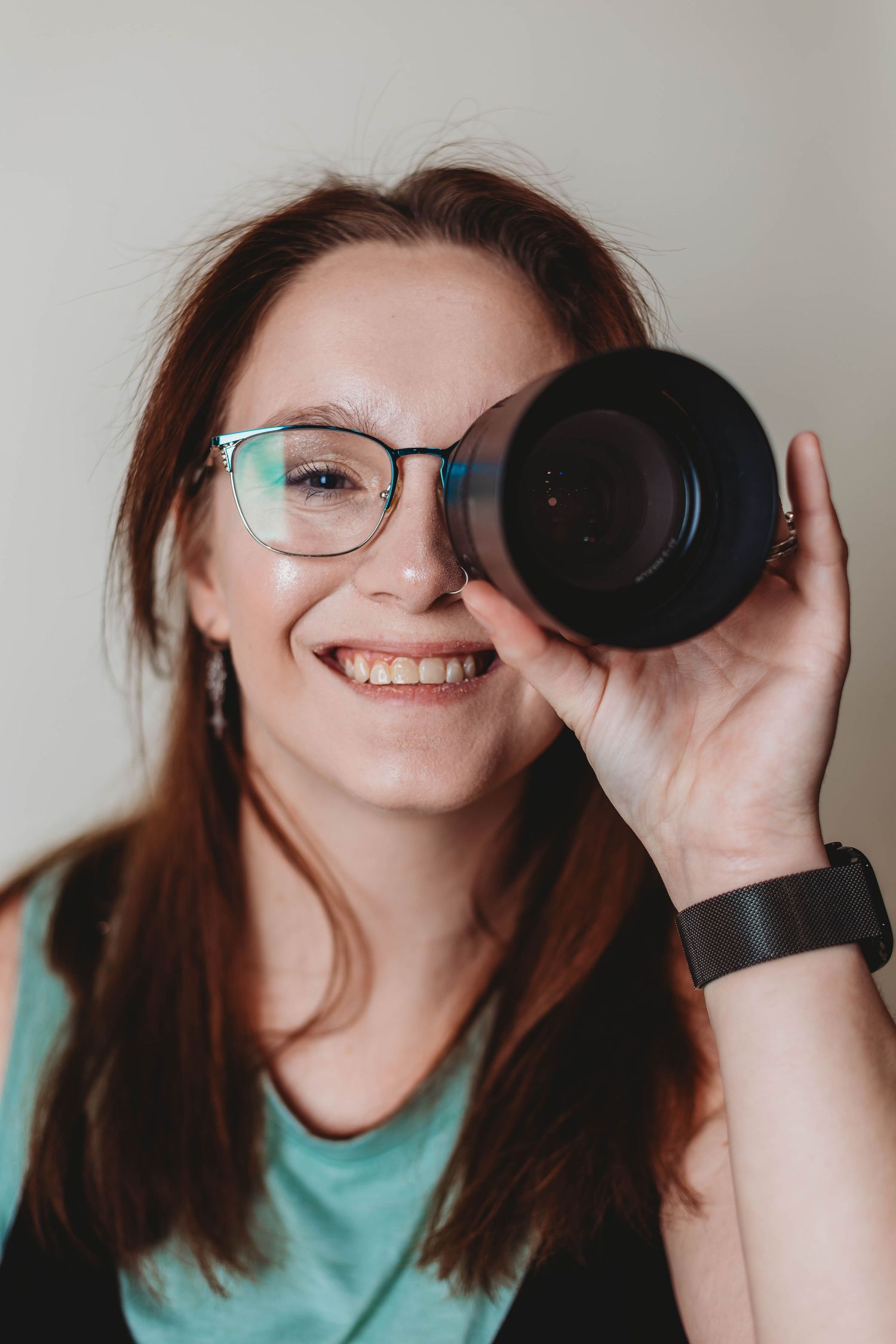 A portrait of someone looking through a camera lens while wearing glasses and a mint colored shirt.
