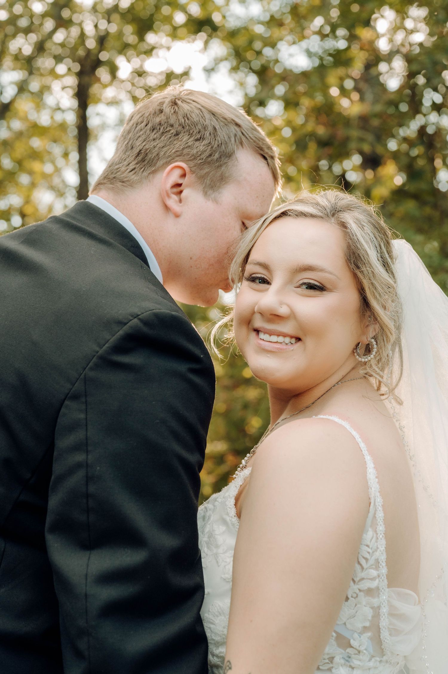 A bride and groom share a romantic moment outdoors with autumn trees in the background.