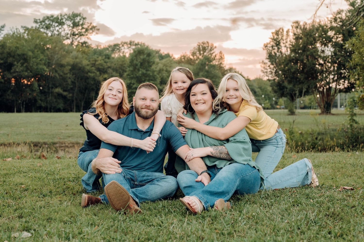 A family in coordinated blue outfits lies together on grass during a sunset photoshoot in a park setting.