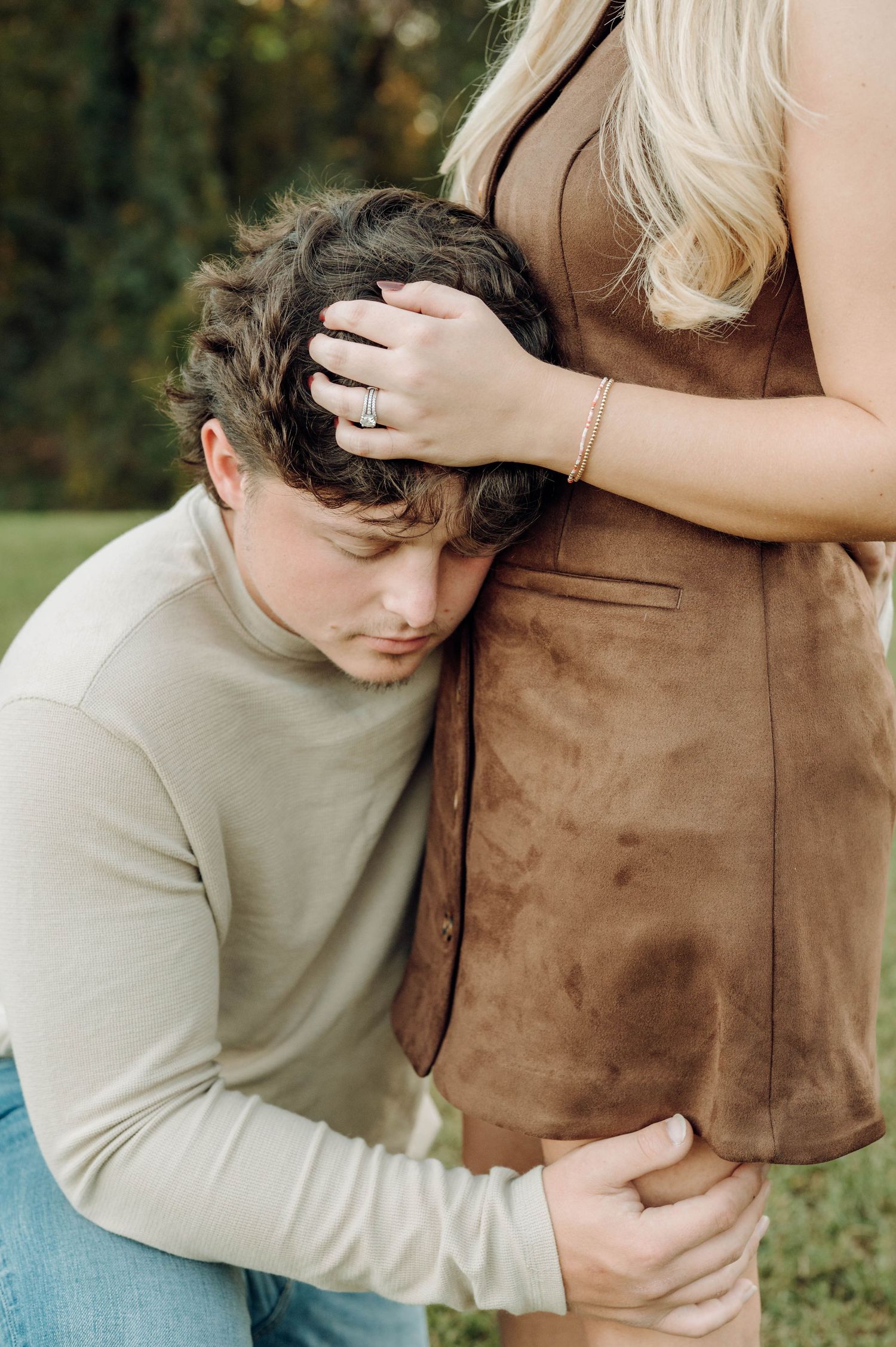 A couple embraces tenderly in an outdoor setting wearing casual attire against a natural backdrop.