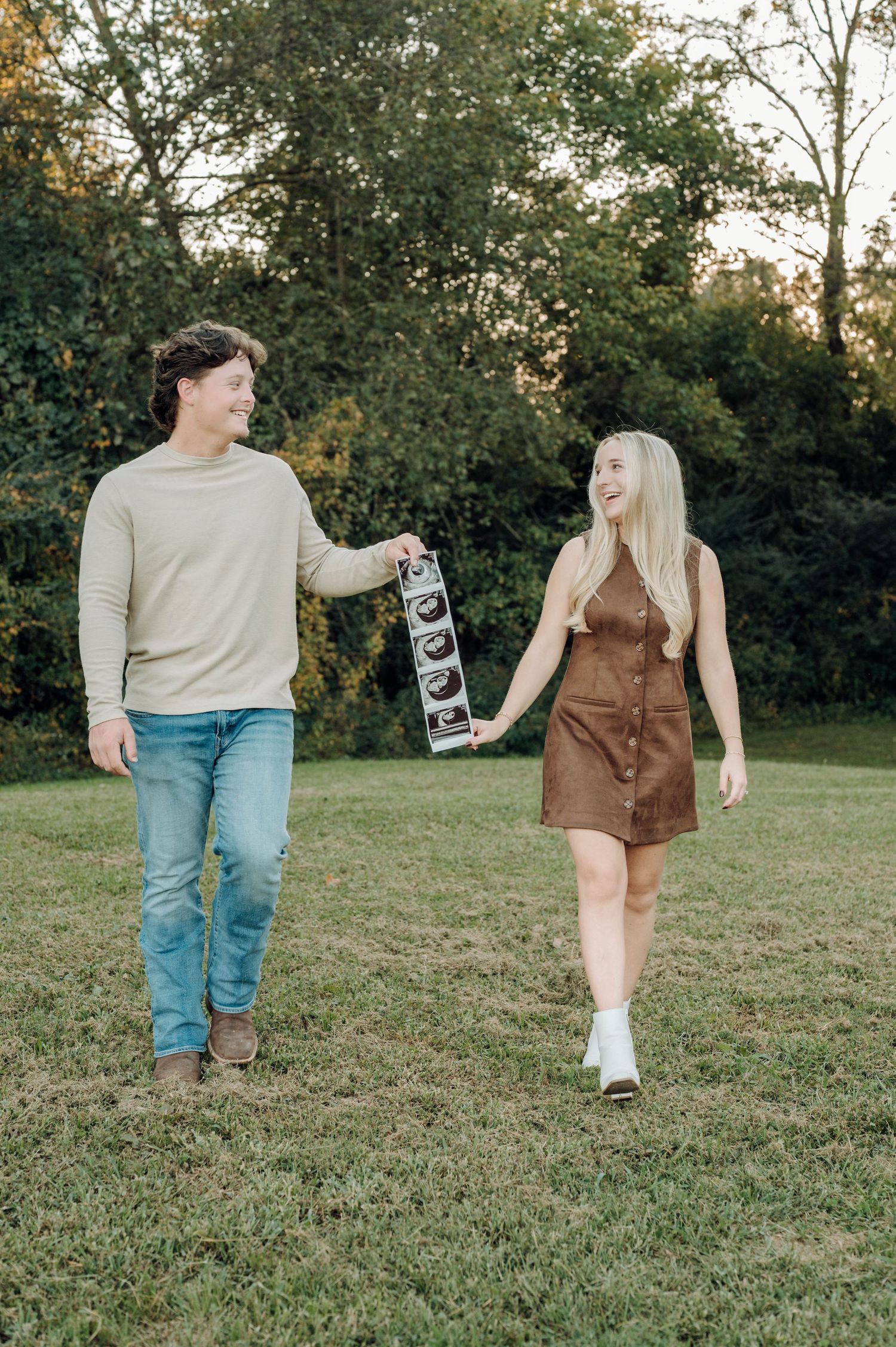 A couple walking together on grass holding ultrasound photos during a pregnancy announcement photoshoot at sunset.
