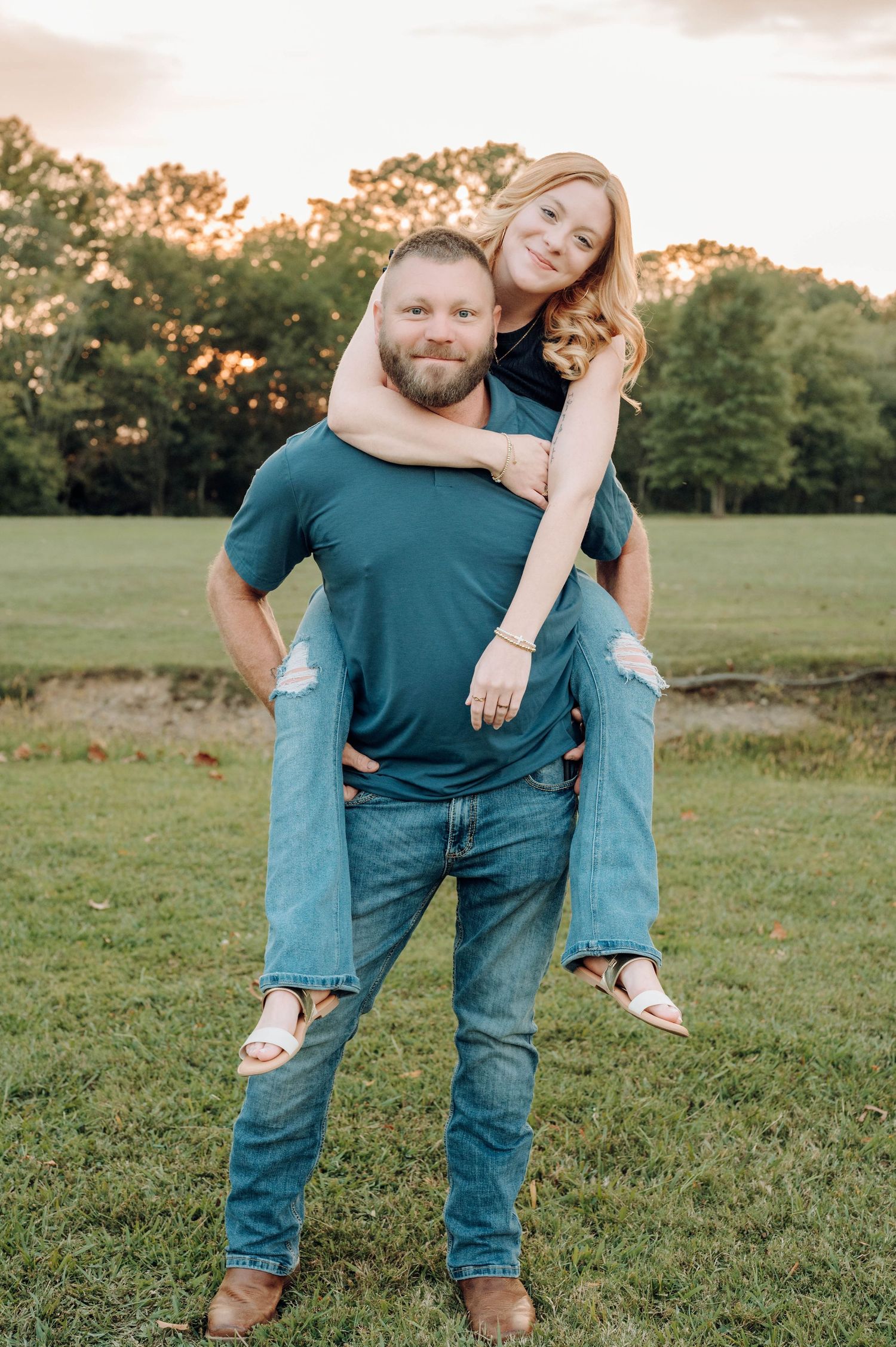 A couple playfully poses in a grassy field at sunset with the person in blue jeans giving a piggyback ride.